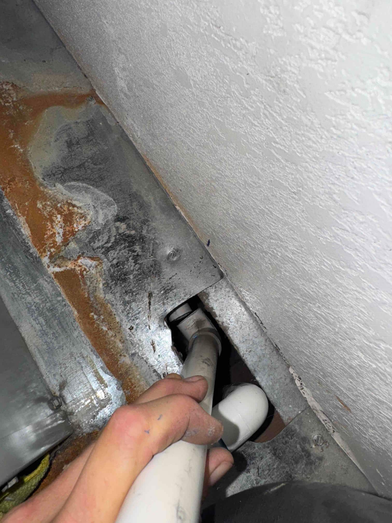 PVC condensate drain piping entering a metal tray near a wall, with visible rust staining.