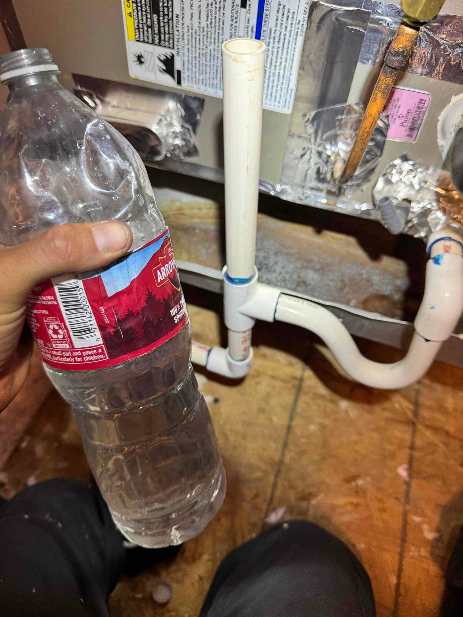 PVC condensate drain with tee and vertical cleanout at the air handler, with a hand holding a water bottle nearby.