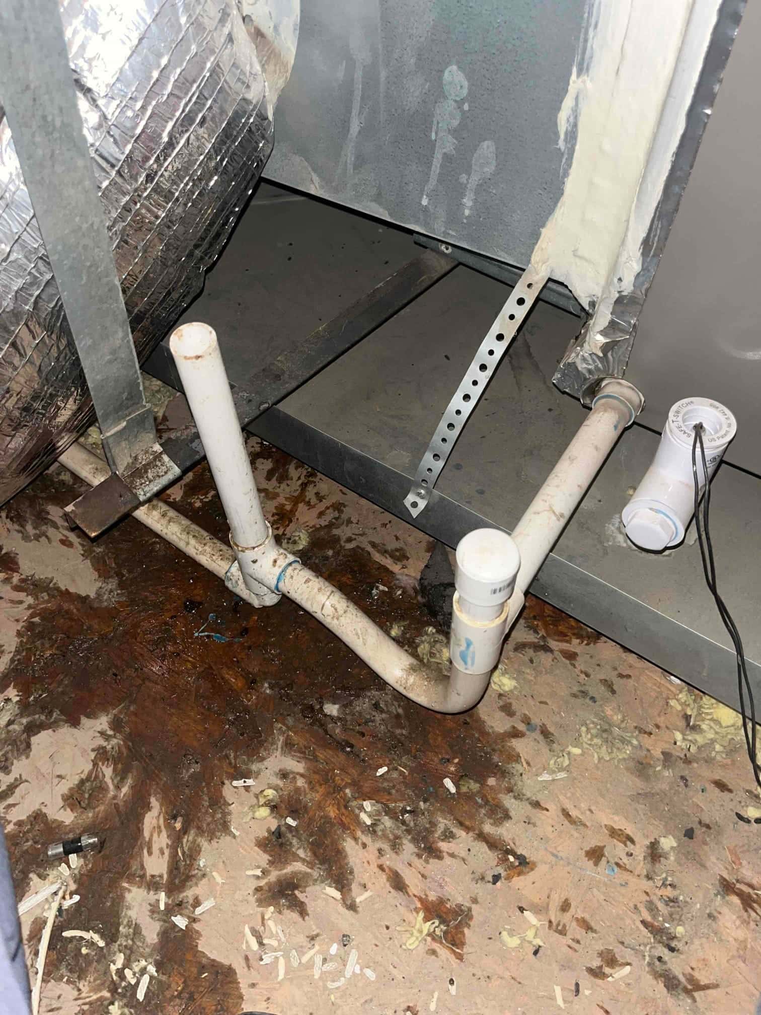 PVC condensate drain with P-trap and cleanout beside air handler, with dark moisture staining on subfloor.