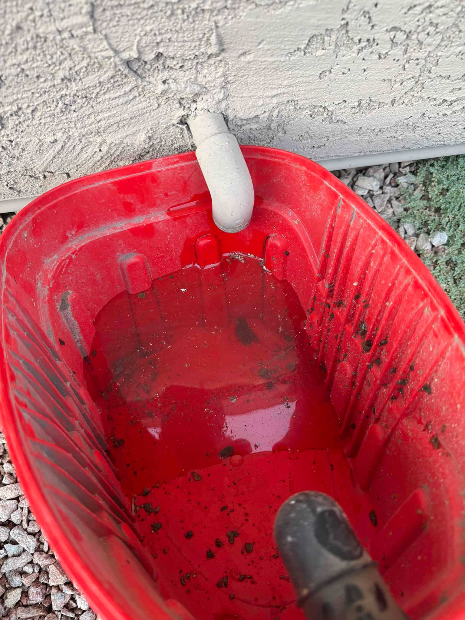 PVC drain outlet at exterior wall dripping into a red bucket with standing water.
