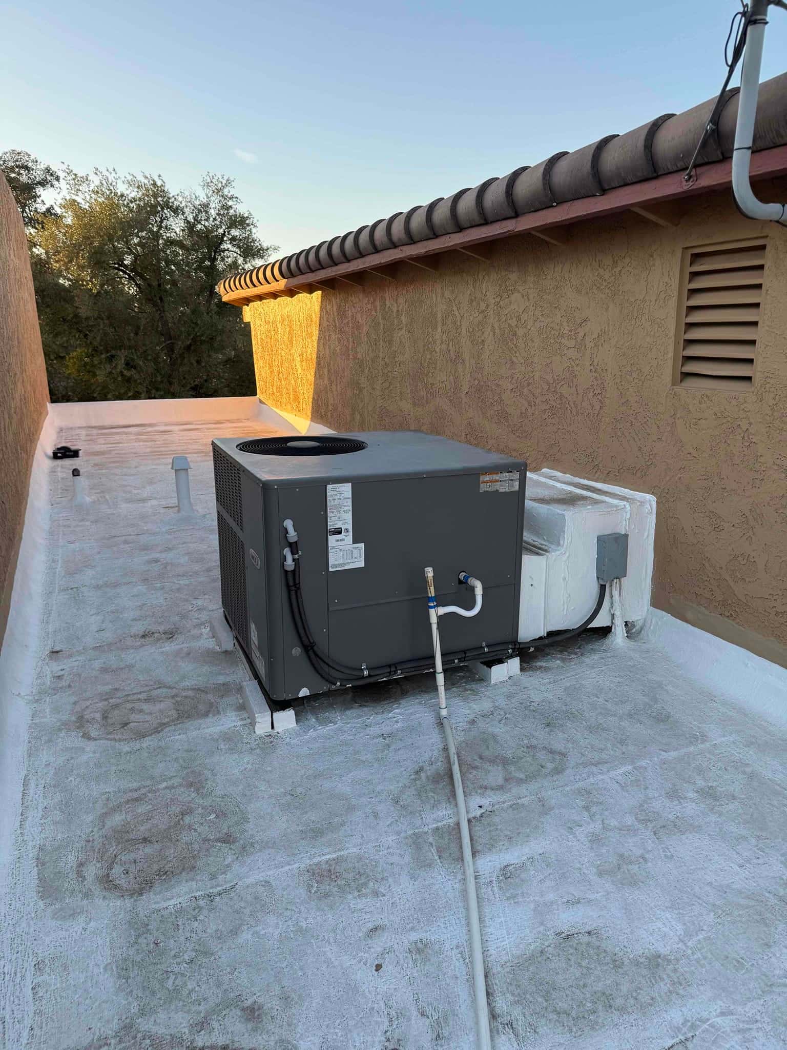 Rooftop air-conditioning unit on flat roof with condensate drain piping, flexible conduits, and nearby service disconnect enclosure.