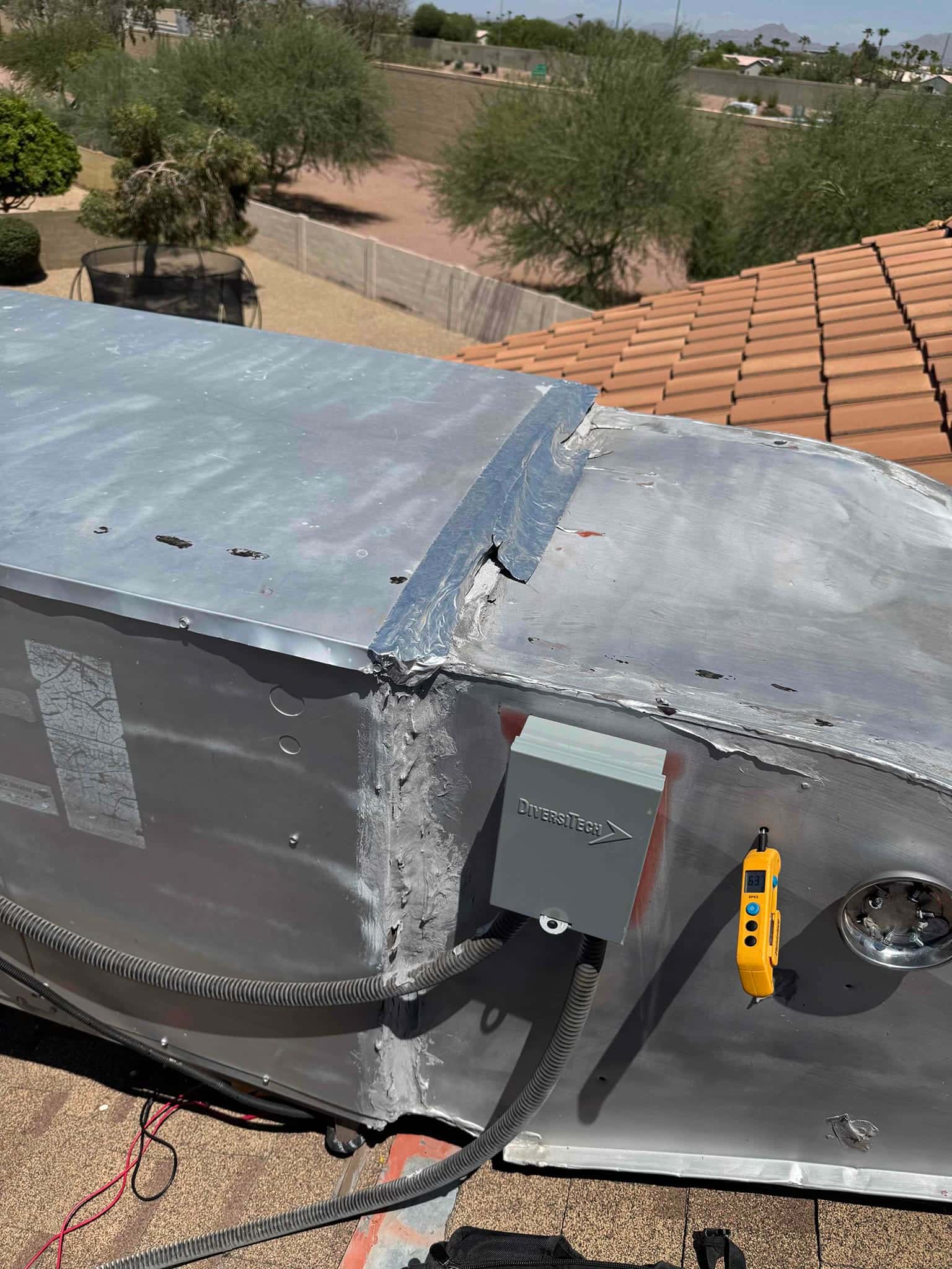 Rooftop HVAC cabinet with sealed seam, service disconnect, and flexible conduits; handheld digital thermometer hangs on the side.
