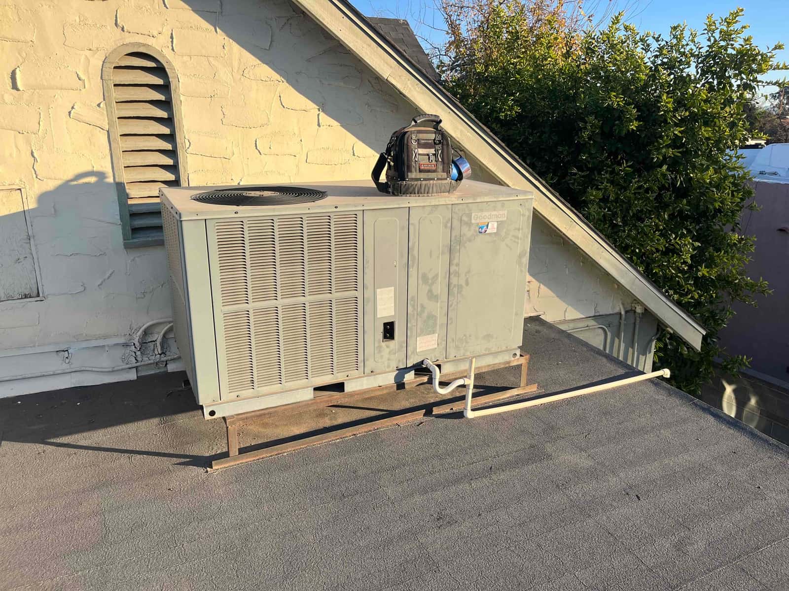 Rooftop packaged HVAC unit on mounting rails with top fan and condensate drain, technician tool bag resting on cabinet.