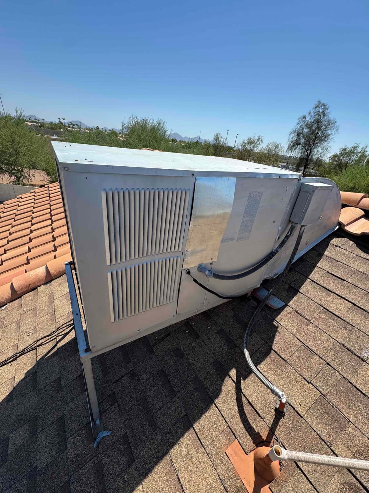 Rooftop packaged HVAC unit on shingle roof with flexible electrical conduit and side louvers visible.