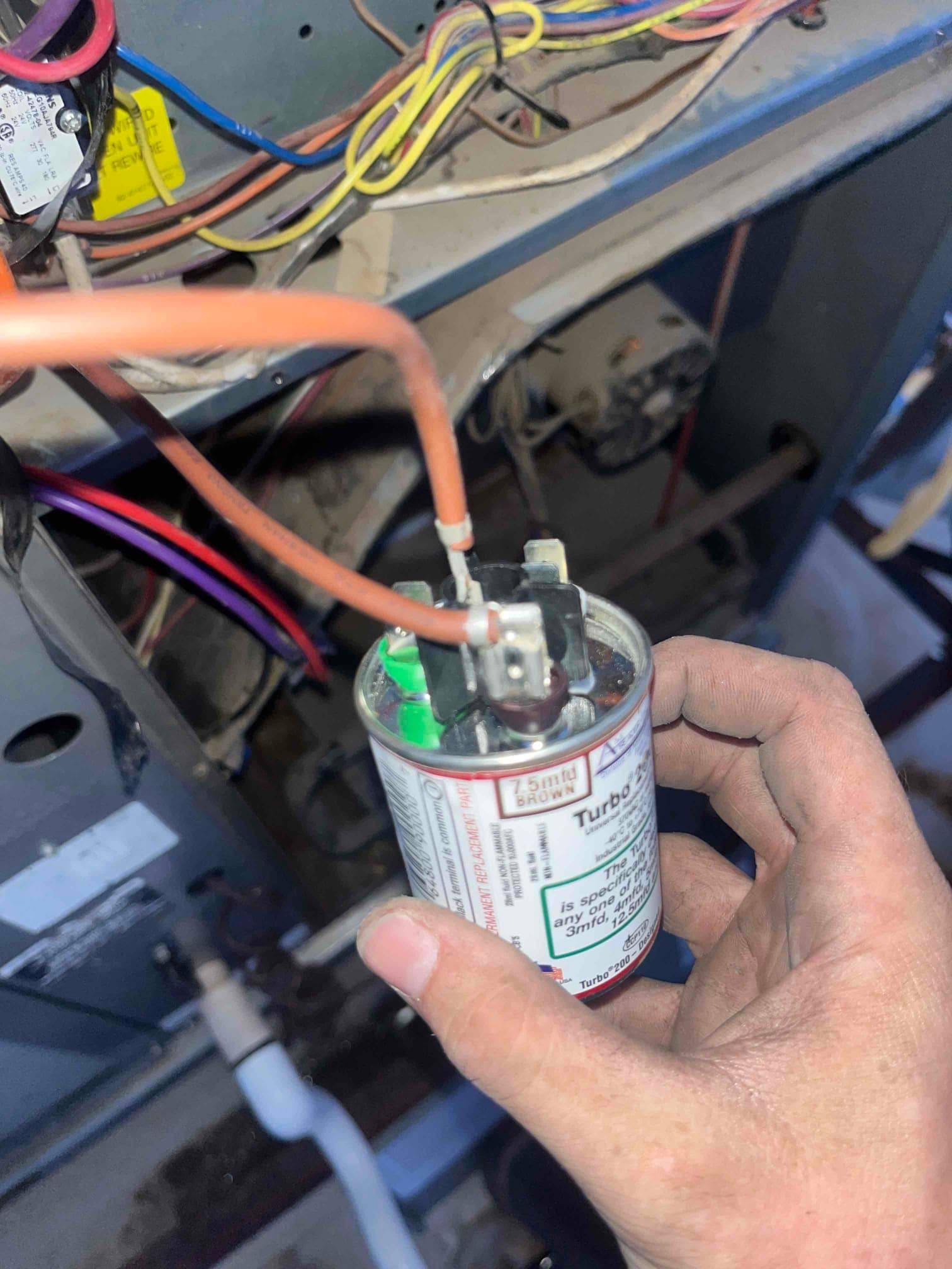 Run capacitor labeled 7.5 mfd BROWN held by hand, with wires attached to its terminals inside the air handler.