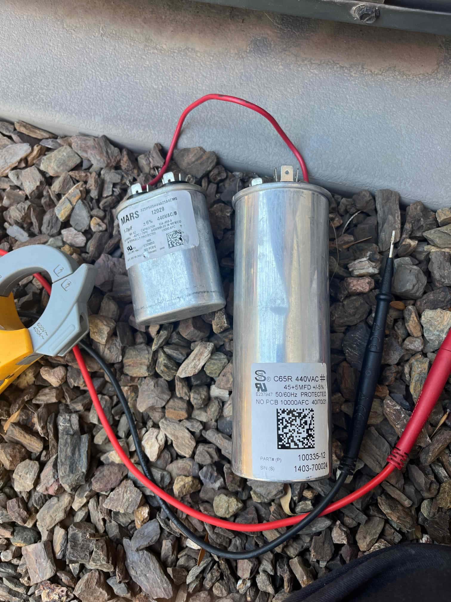 Two run capacitors on the ground with meter leads; one labeled 45+5 μF 440VAC, another labeled 5.0 μF 440VAC.