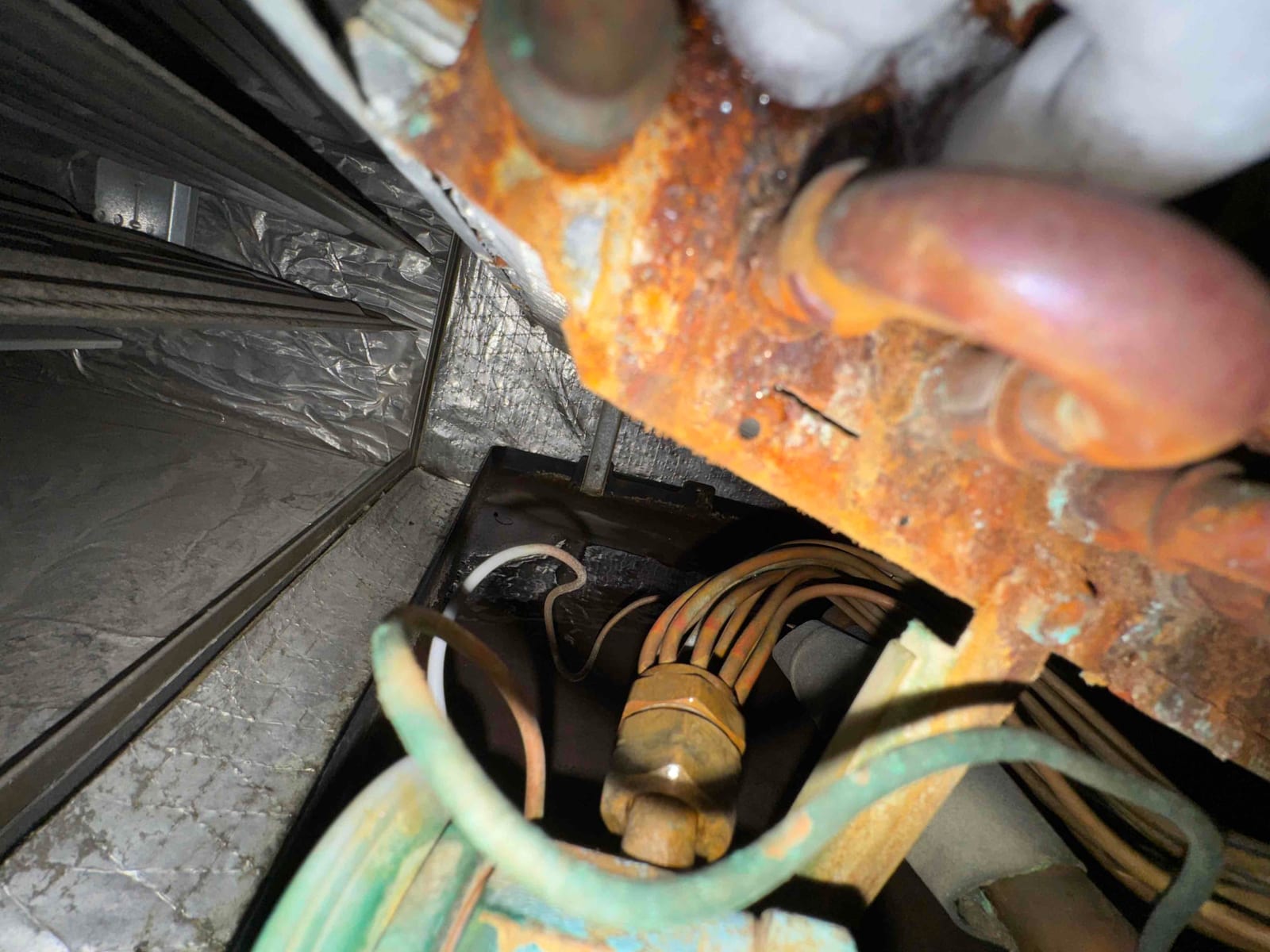 Rusted metal panel with copper refrigerant lines and wiring harness near the evaporator coil fins inside the air handler.