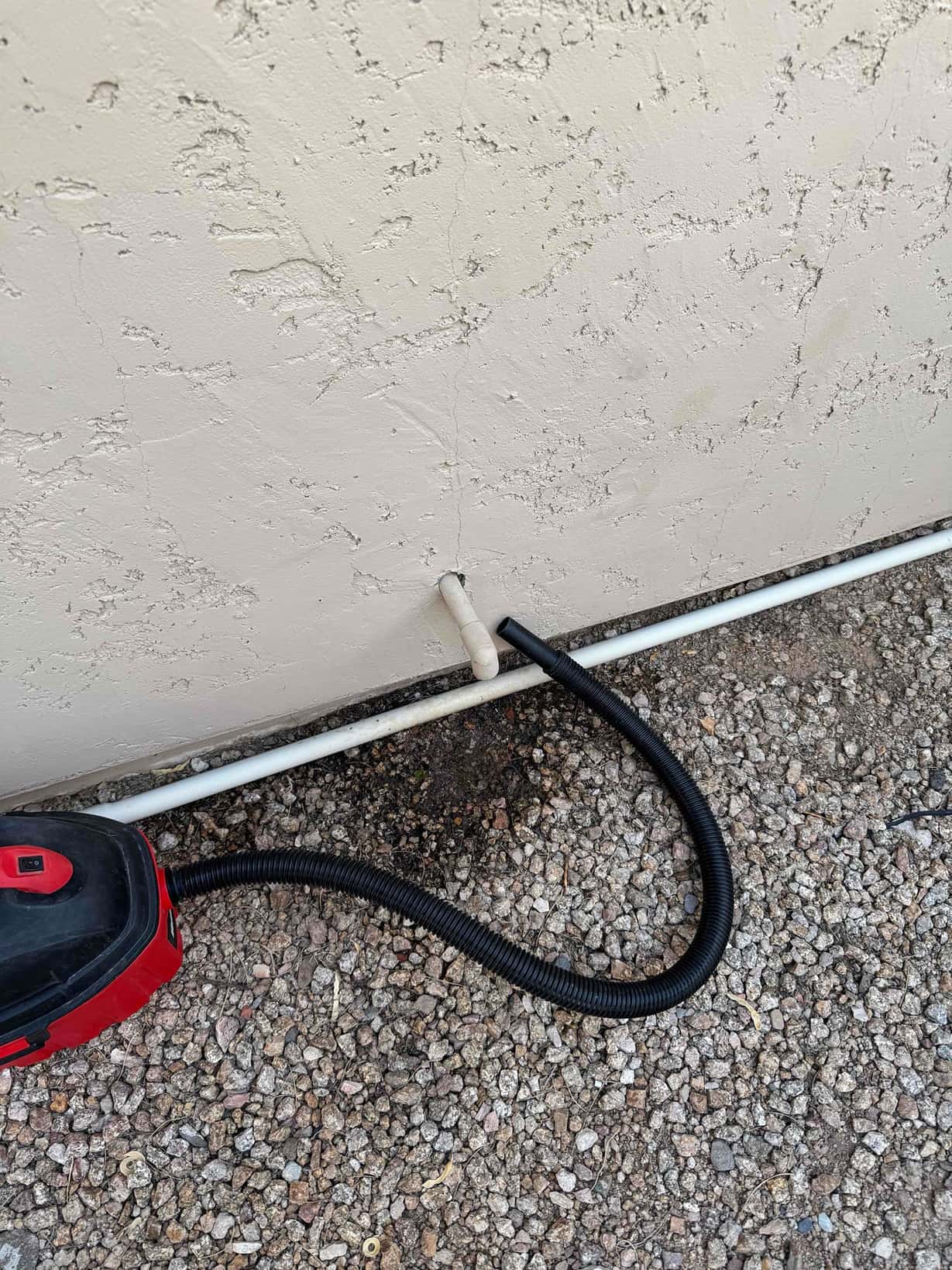 Shop vacuum hose attached to an exterior condensate drain line at the wall over gravel.