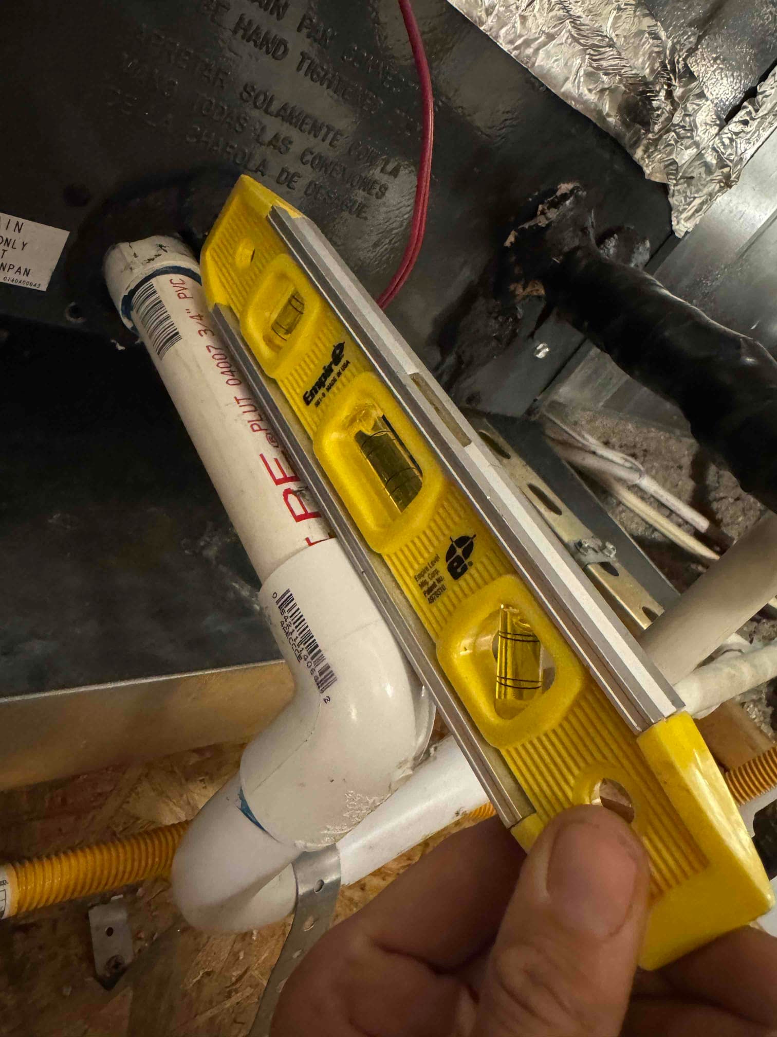 Small bubble level held against PVC condensate drain near air handler, with insulated refrigerant line entering cabinet and sealant applied.