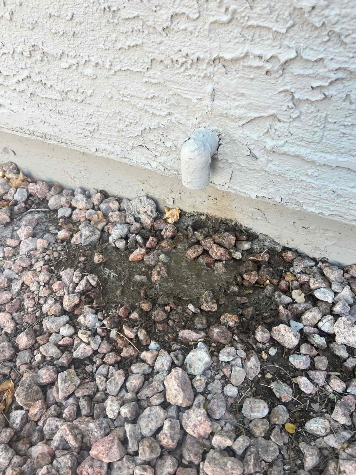 Small PVC elbow drain outlet on exterior wall, with wet stones directly below the opening.