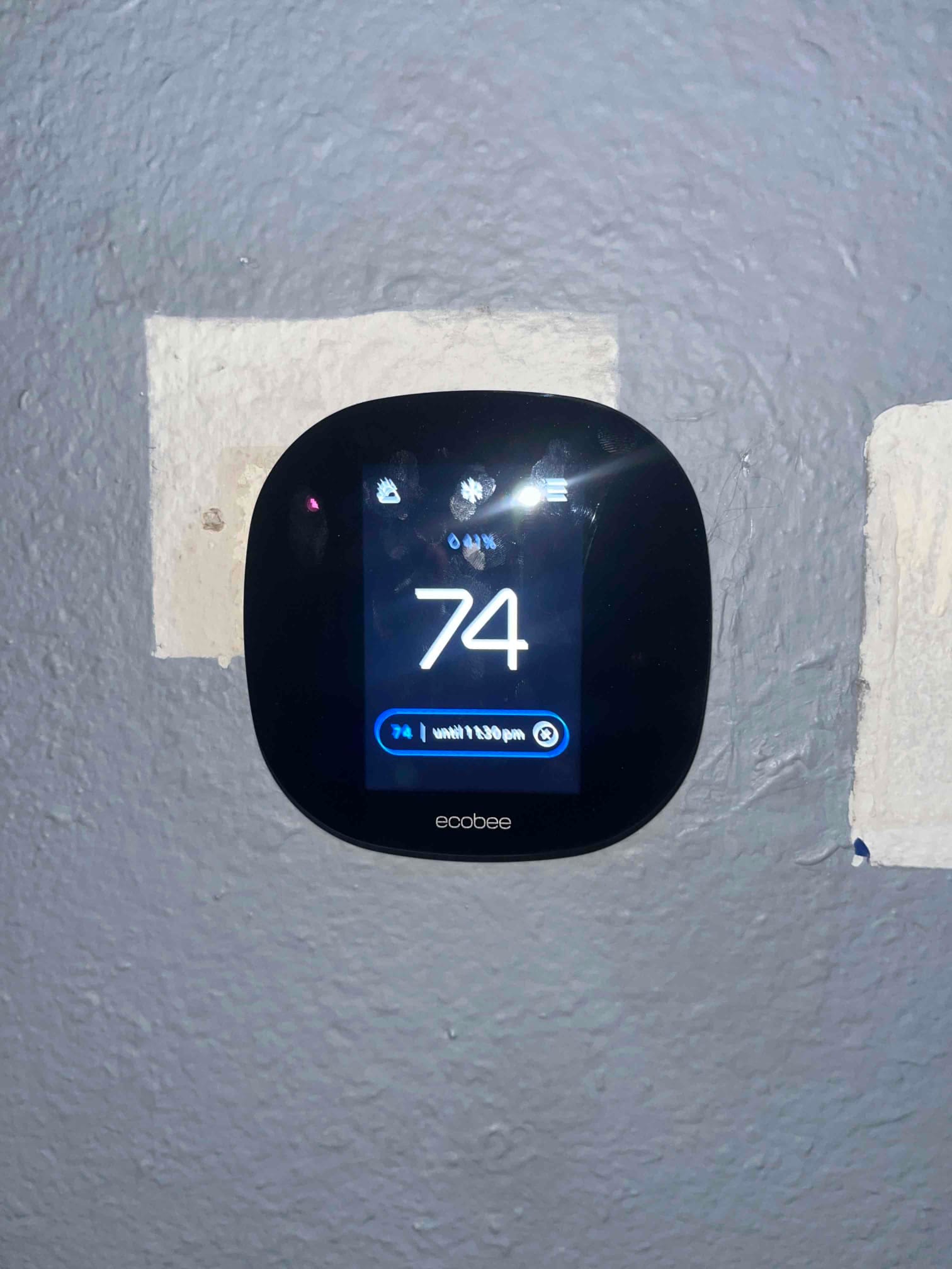 Smart thermostat on interior wall displays 74 degrees with cooling mode icon and a hold set until 1:30 pm.