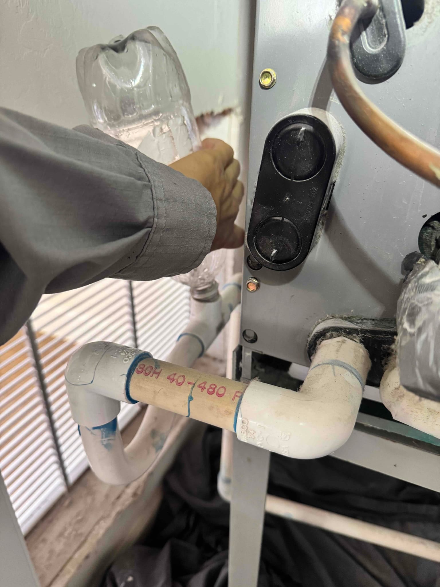 Technician hand holds a plastic bottle near condensate drain PVC at indoor air handler cabinet.