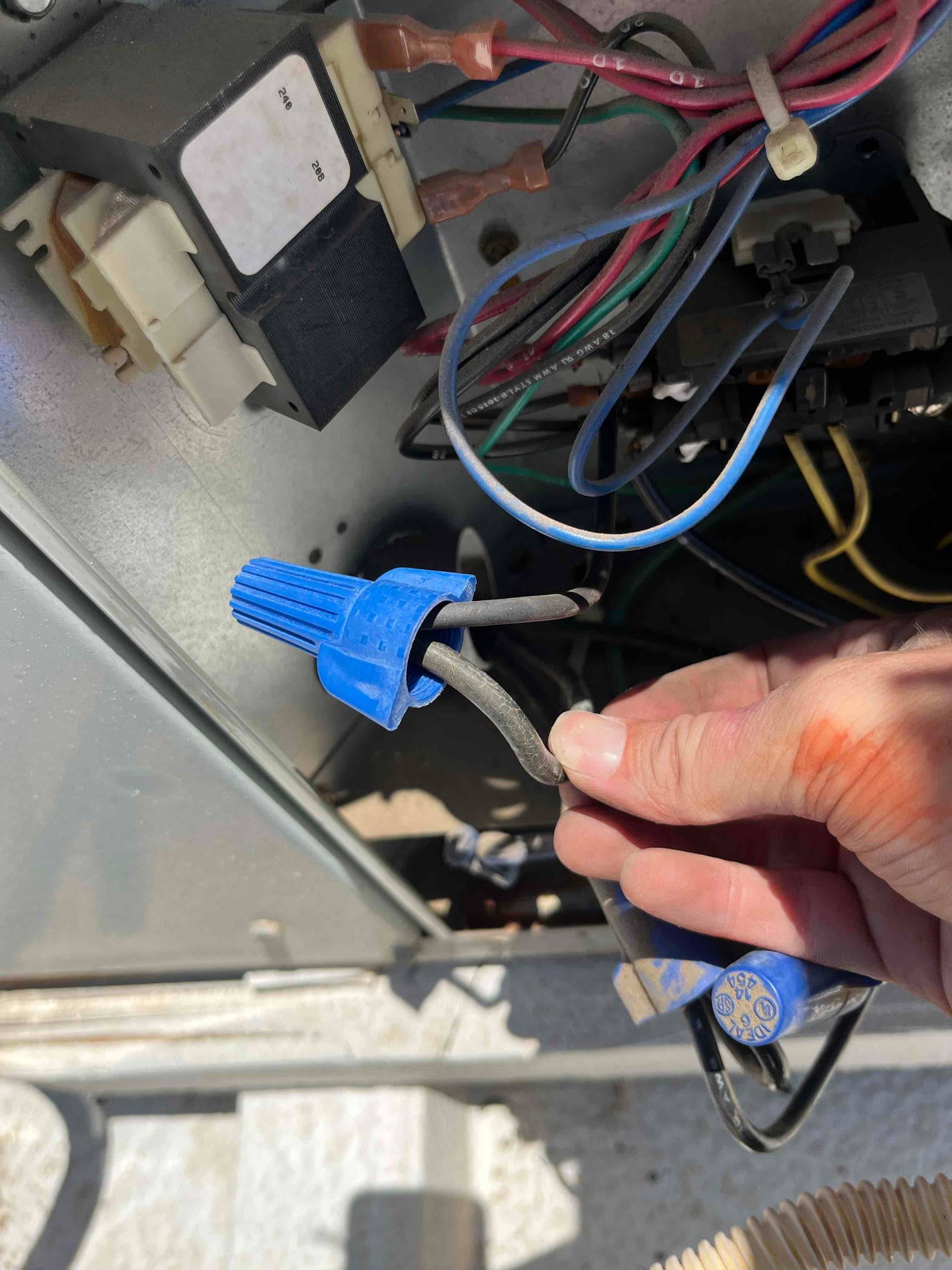 Technician holds a blue wire connector with two black conductors inside an outdoor condenser control compartment.