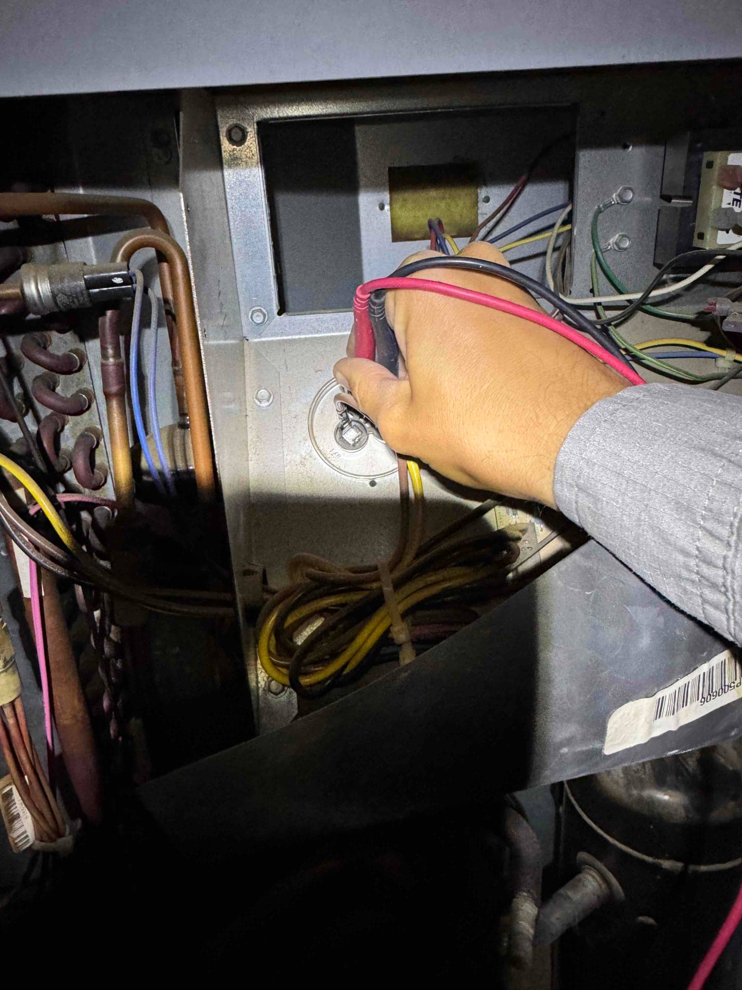 Technician holds meter leads inside the outdoor condenser compartment, showing wiring harnesses, copper refrigerant lines, and the compressor in the background.