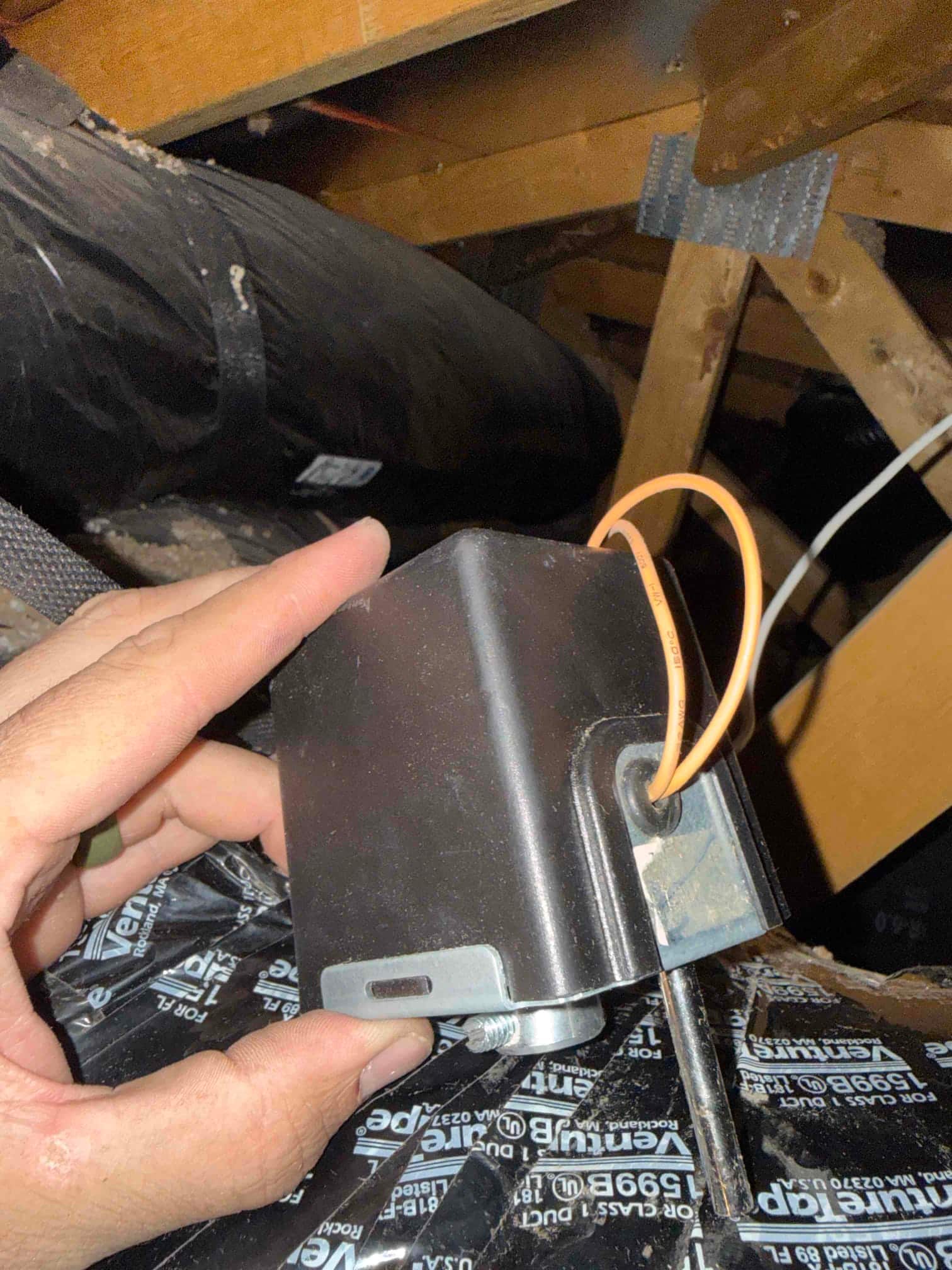 Technician holds a small black electrical housing with orange wires in an attic near ductwork.