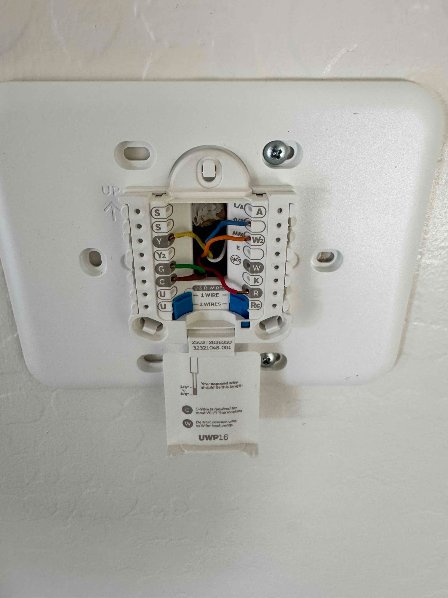 Thermostat wall plate with colored control wires secured to labeled terminals, mounting screws installed, and instruction tag extended.