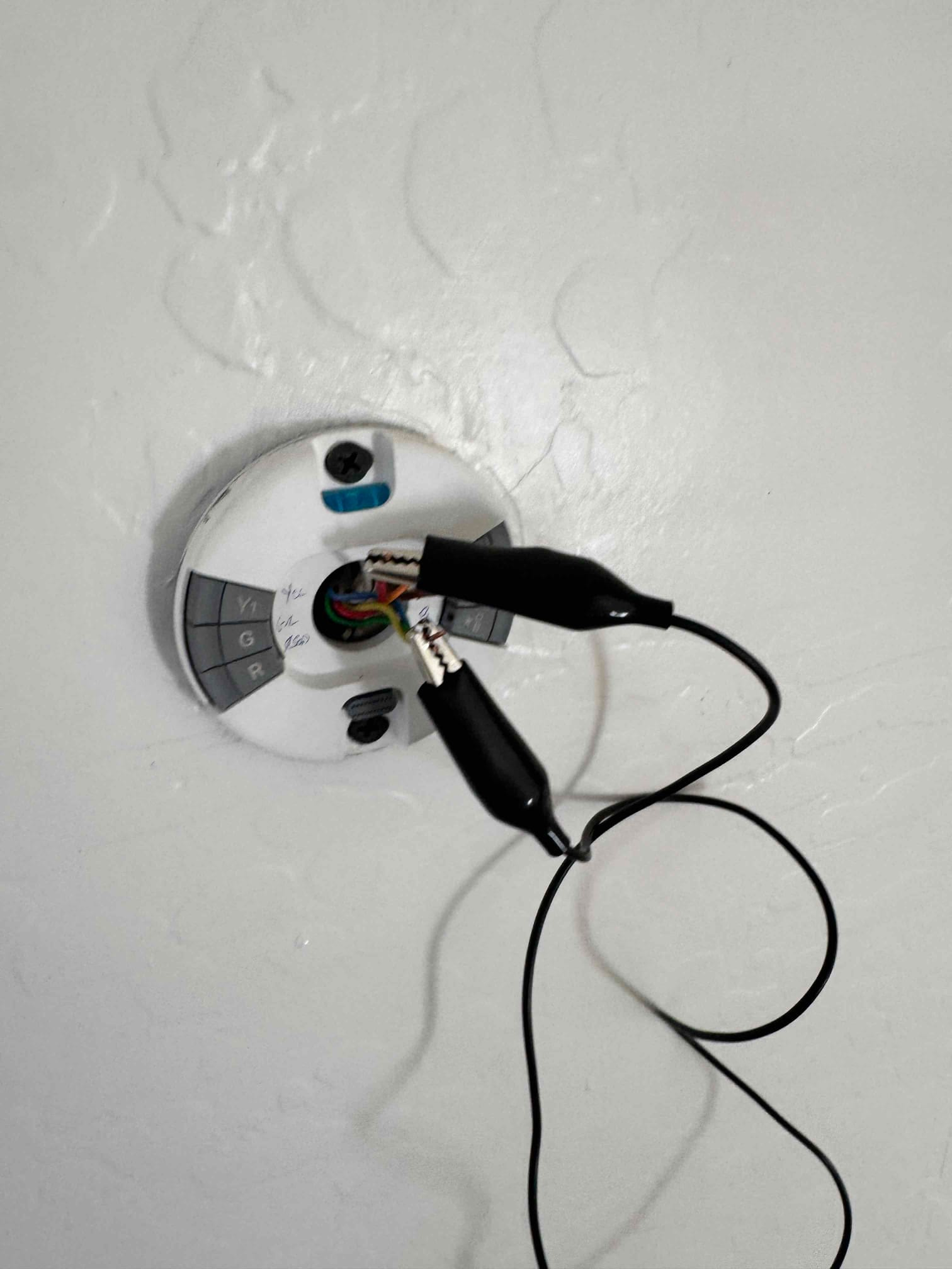 Thermostat wall plate with exposed low-voltage wires and two alligator clip leads connected for testing.
