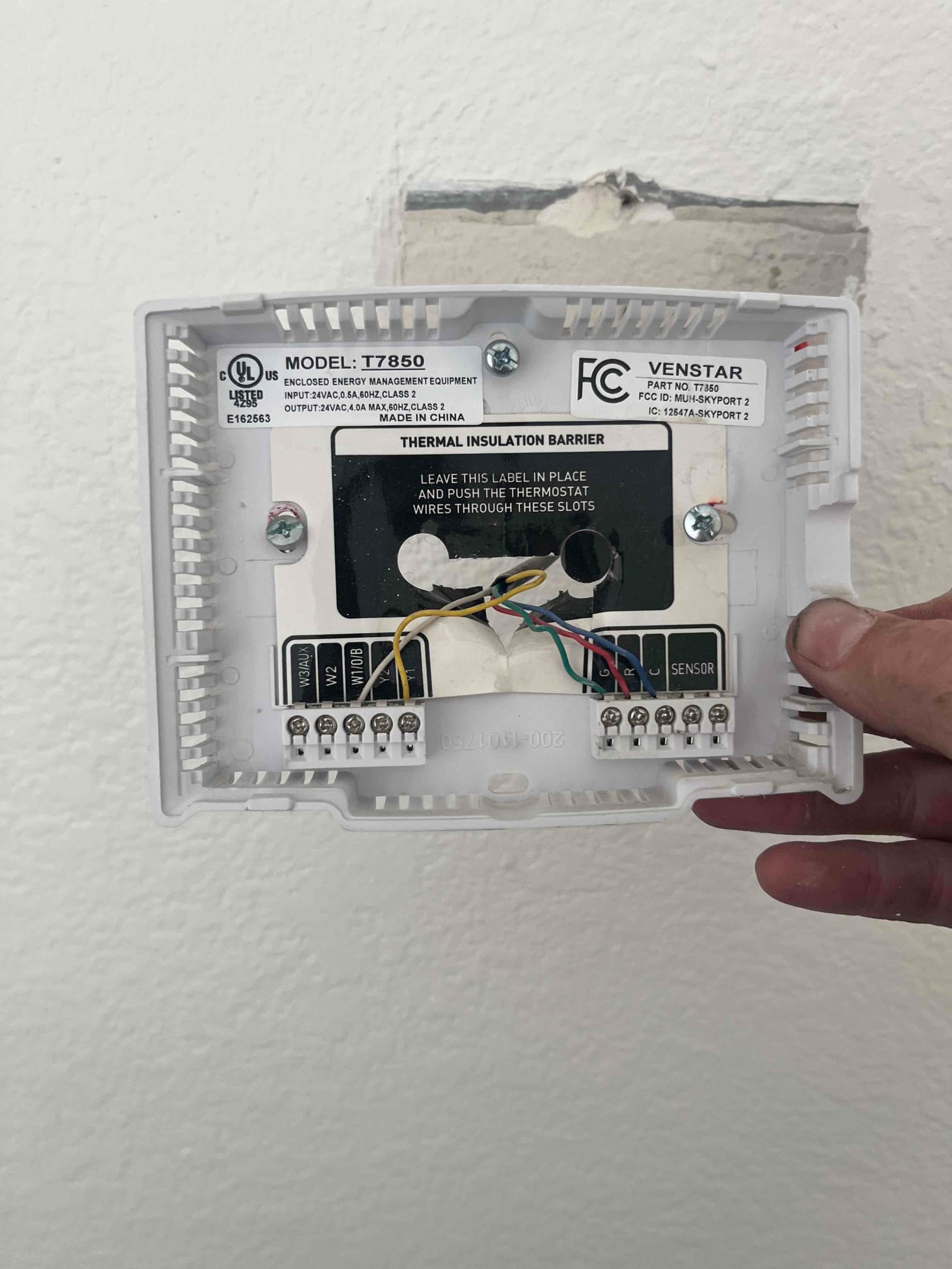 Thermostat wall plate removed, showing low-voltage control wires connected to labeled terminals and an open wall cavity.