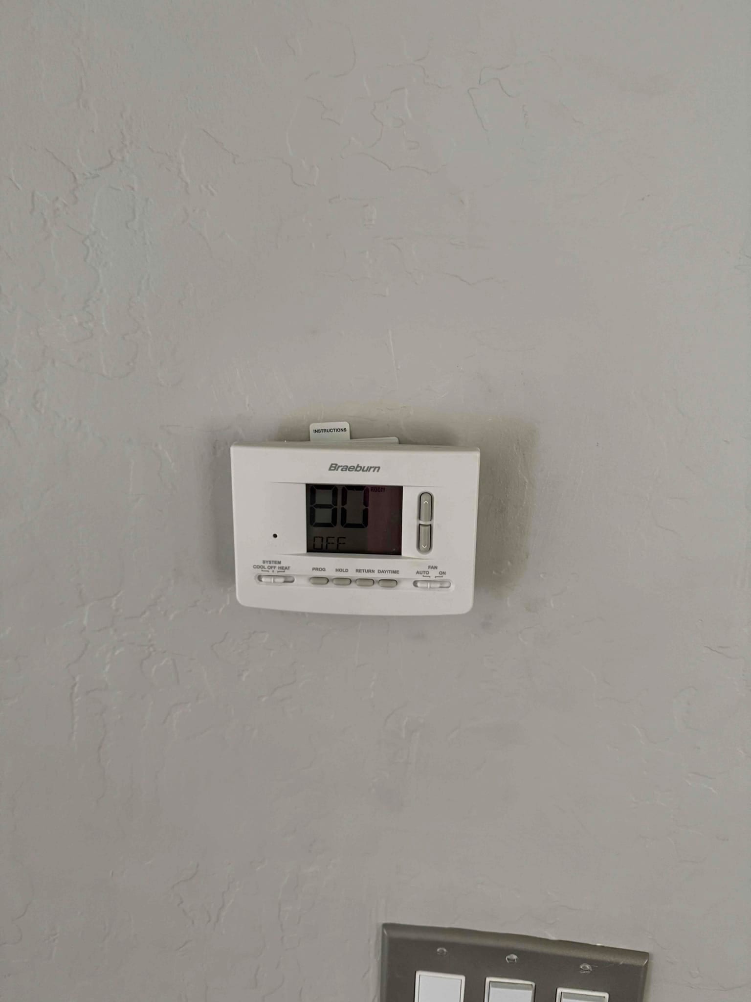 Wall-mounted digital thermostat displaying 80 with system set to OFF; control buttons and instruction card visible.