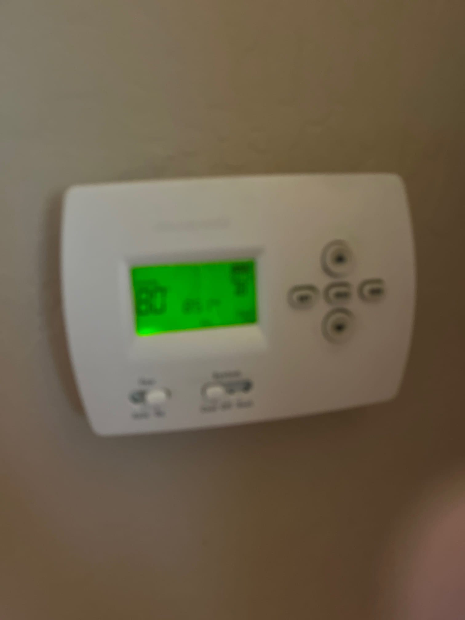 Wall-mounted digital thermostat with green backlit screen and navigation buttons, with fan and system mode switches below.