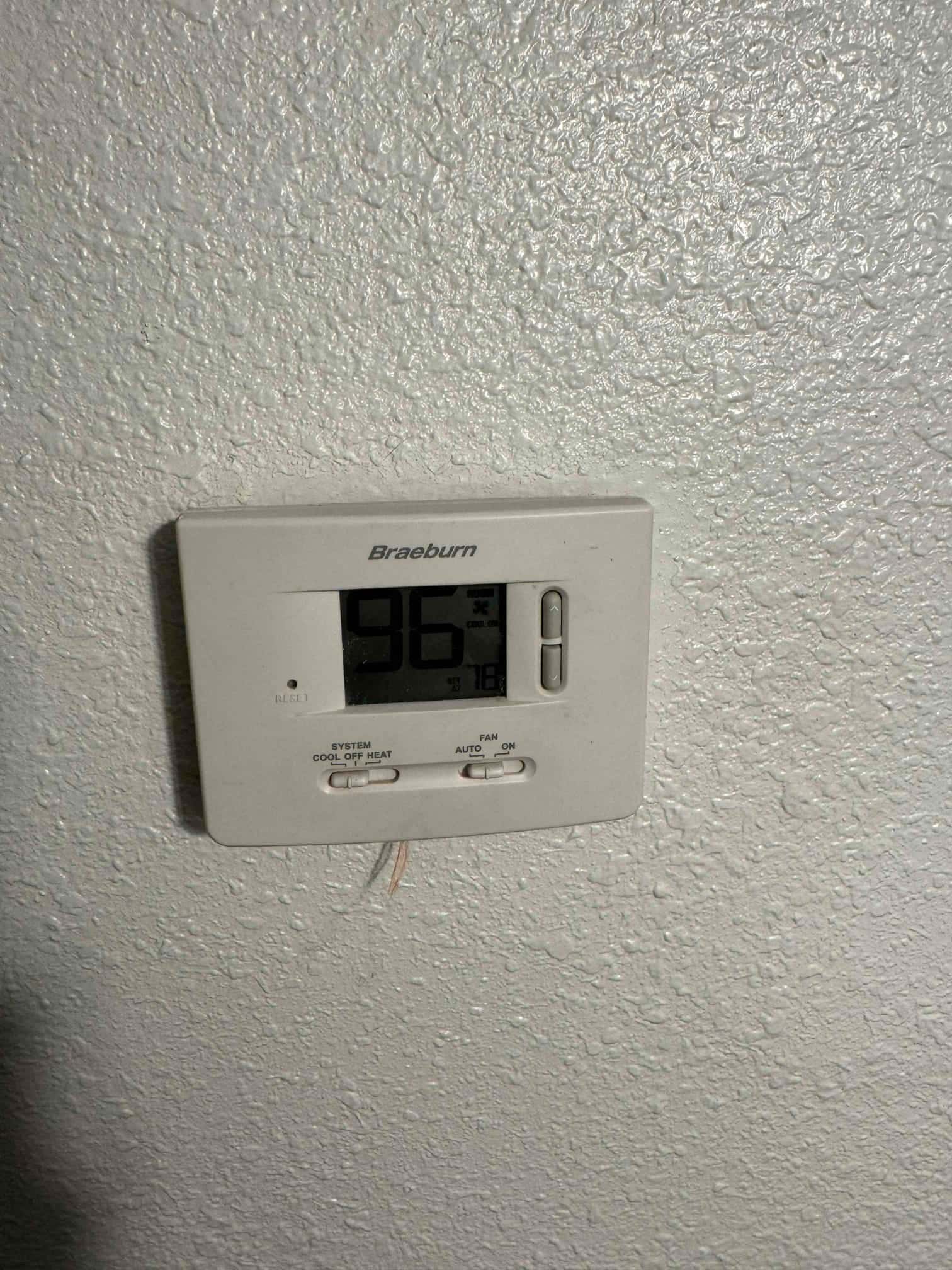 Wall-mounted digital thermostat shows 96 on the screen, with system and fan sliders and a protruding wire.