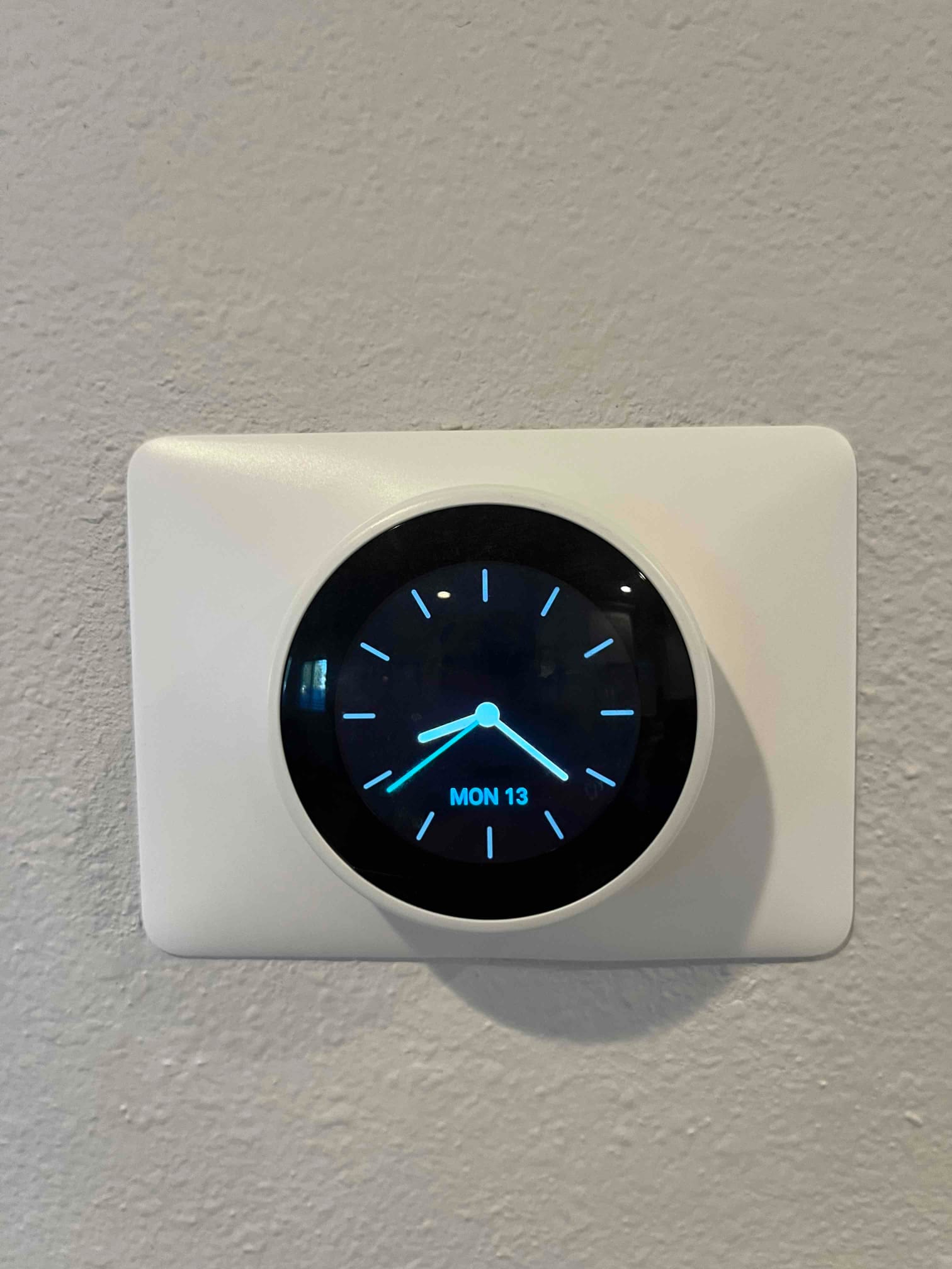 Wall-mounted smart thermostat displays an analog clock screen with day and date shown as MON 13.