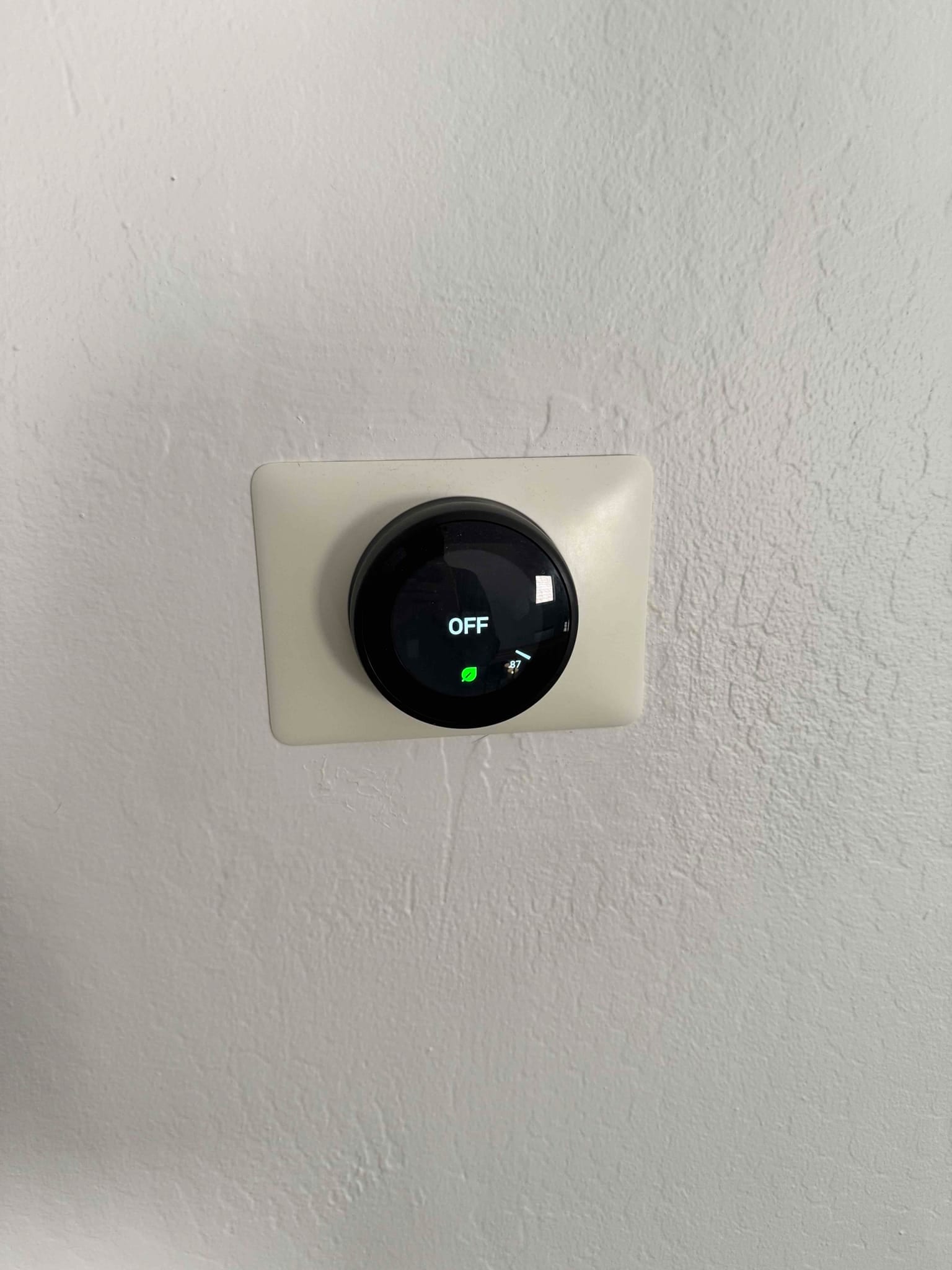 Wall-mounted smart thermostat displays OFF with a green leaf icon on a square trim plate.