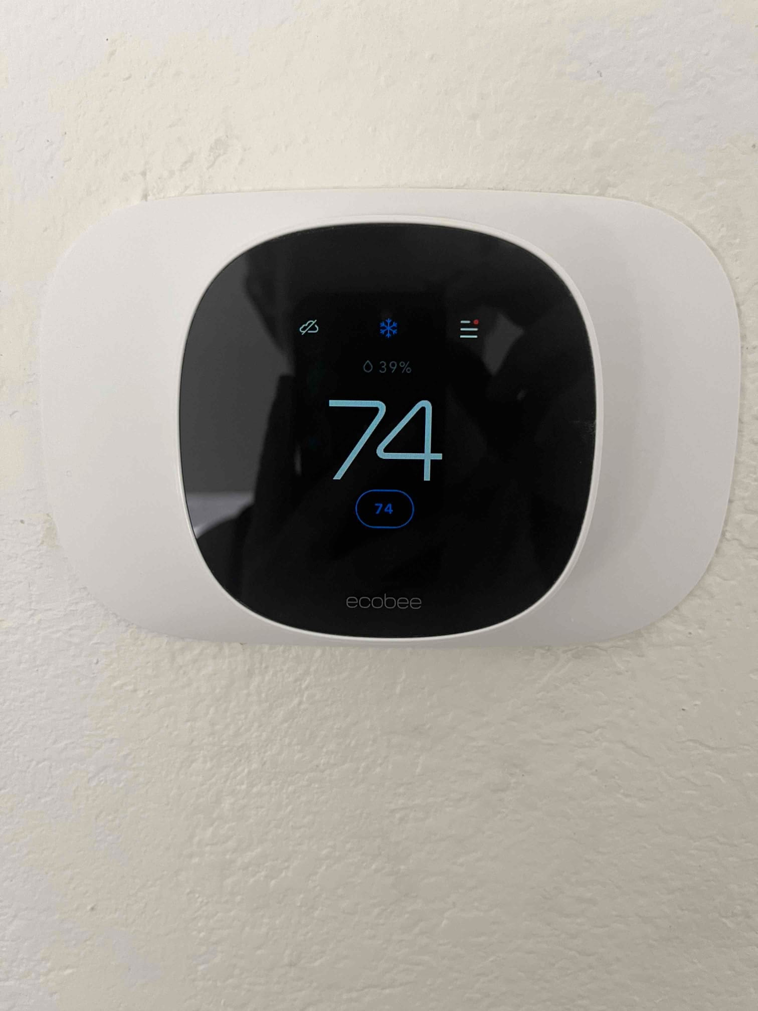 Wall-mounted smart thermostat shows cooling mode active, screen displaying 74 and 39% humidity on a textured wall.