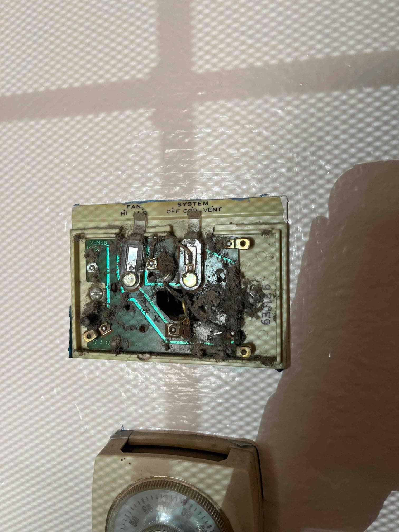 Wall-mounted thermostat backplate with heavy dust buildup, charred circuit board area, and exposed terminals labeled fan and system.