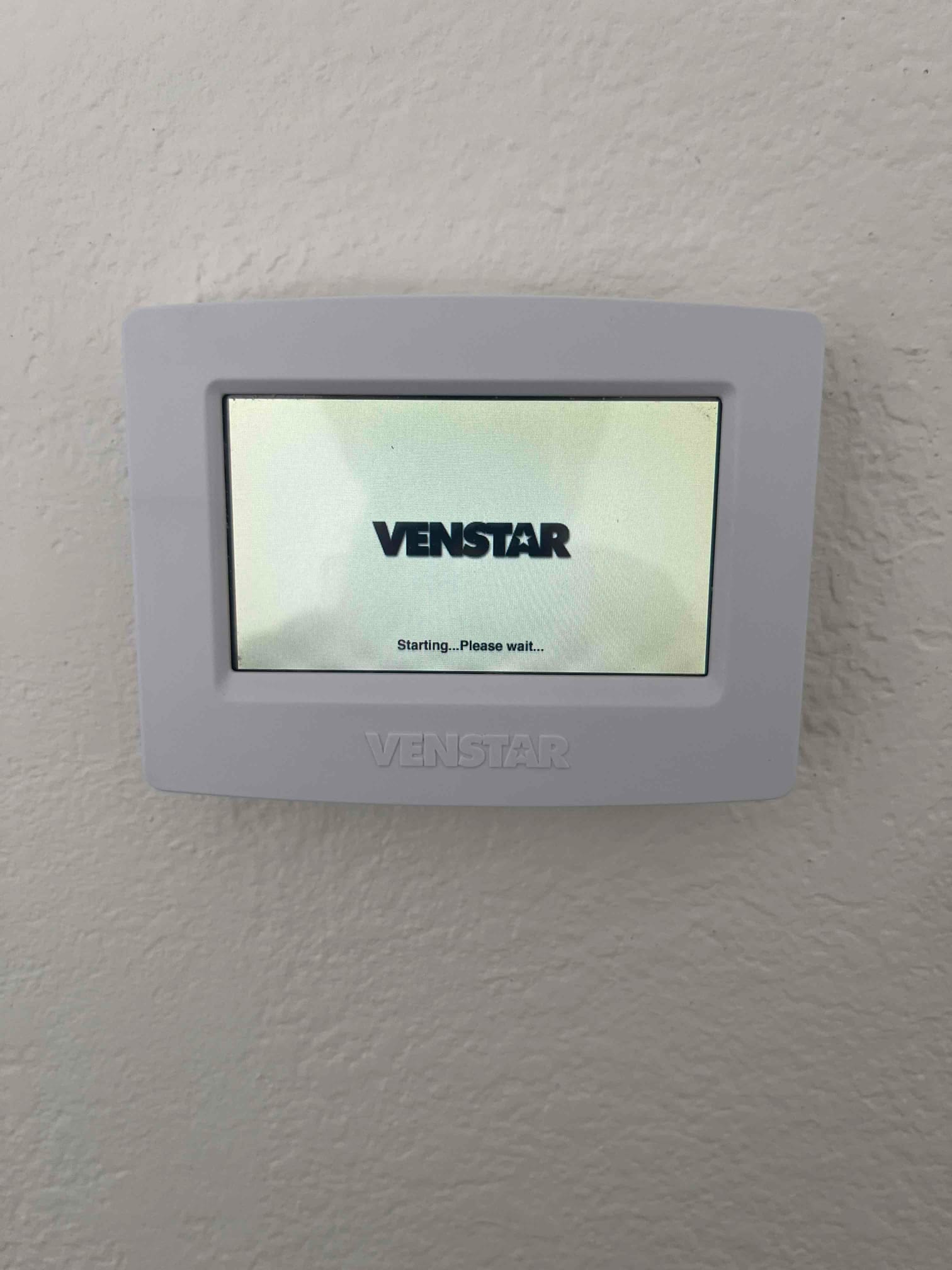 Wall-mounted touchscreen thermostat displays a startup screen with the message 'Starting... Please wait...' on its display.