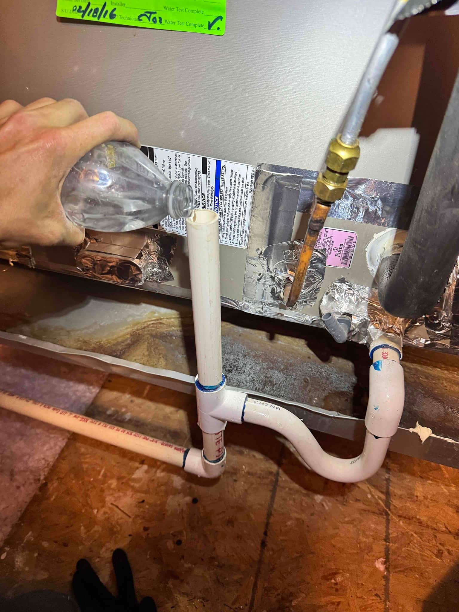 Water pours into the air handler’s condensate drain cleanout, showing PVC trap assembly and nearby insulated refrigerant lines.