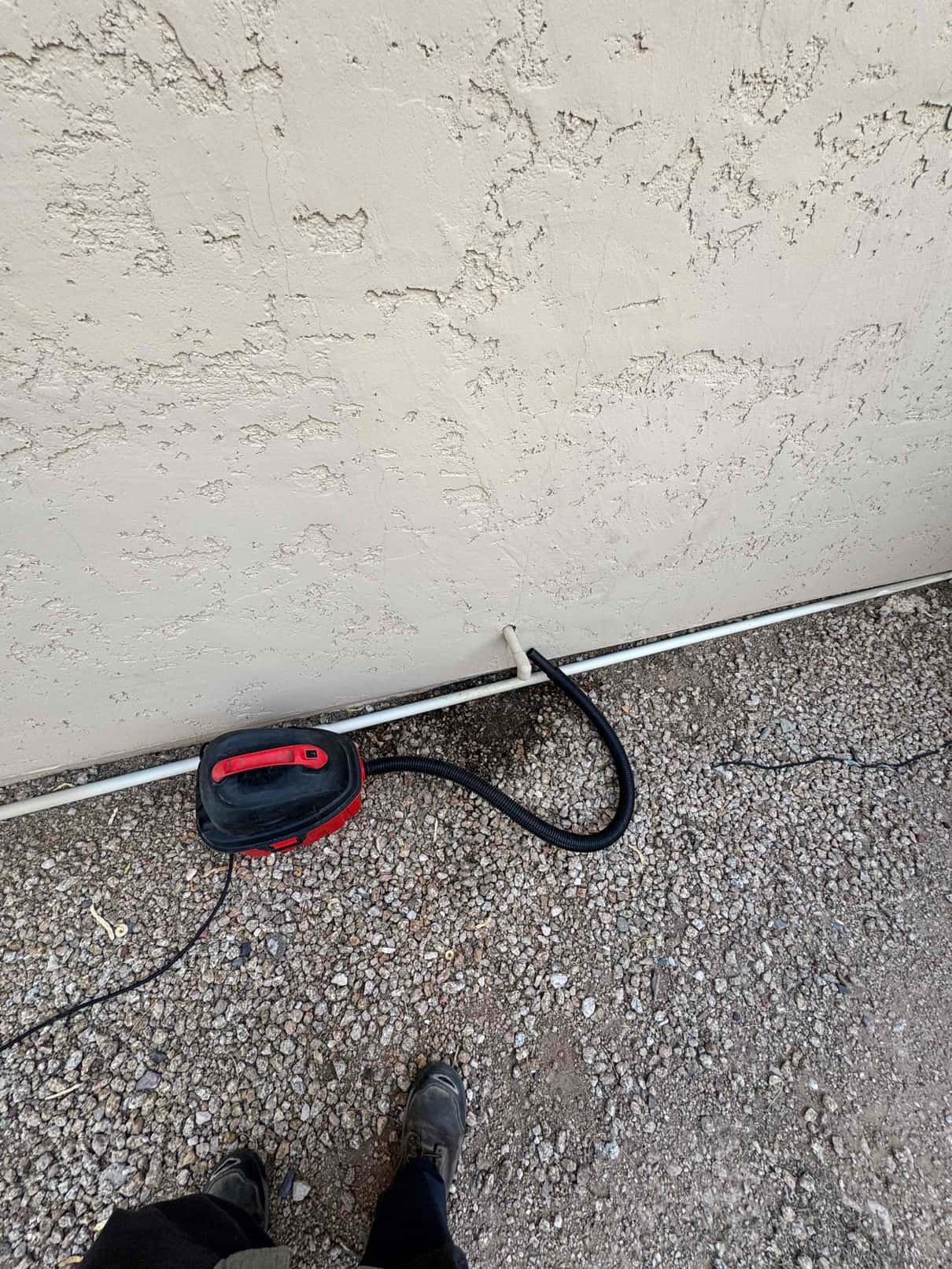 Wet/dry vacuum hose connected to a small PVC drain line exiting the exterior wall over gravel.