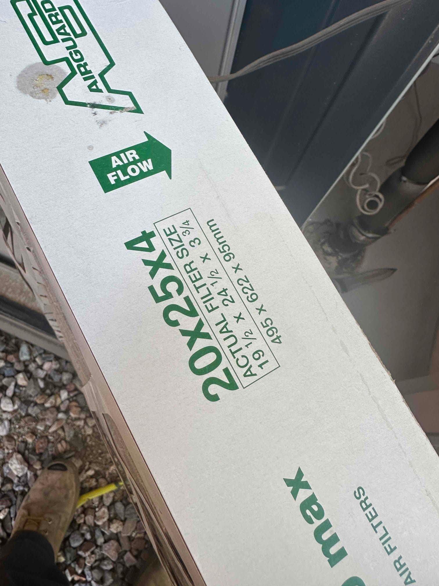 Air filter box labeled 20x25x4 with airflow arrow, resting on gravel beside metal equipment housing.