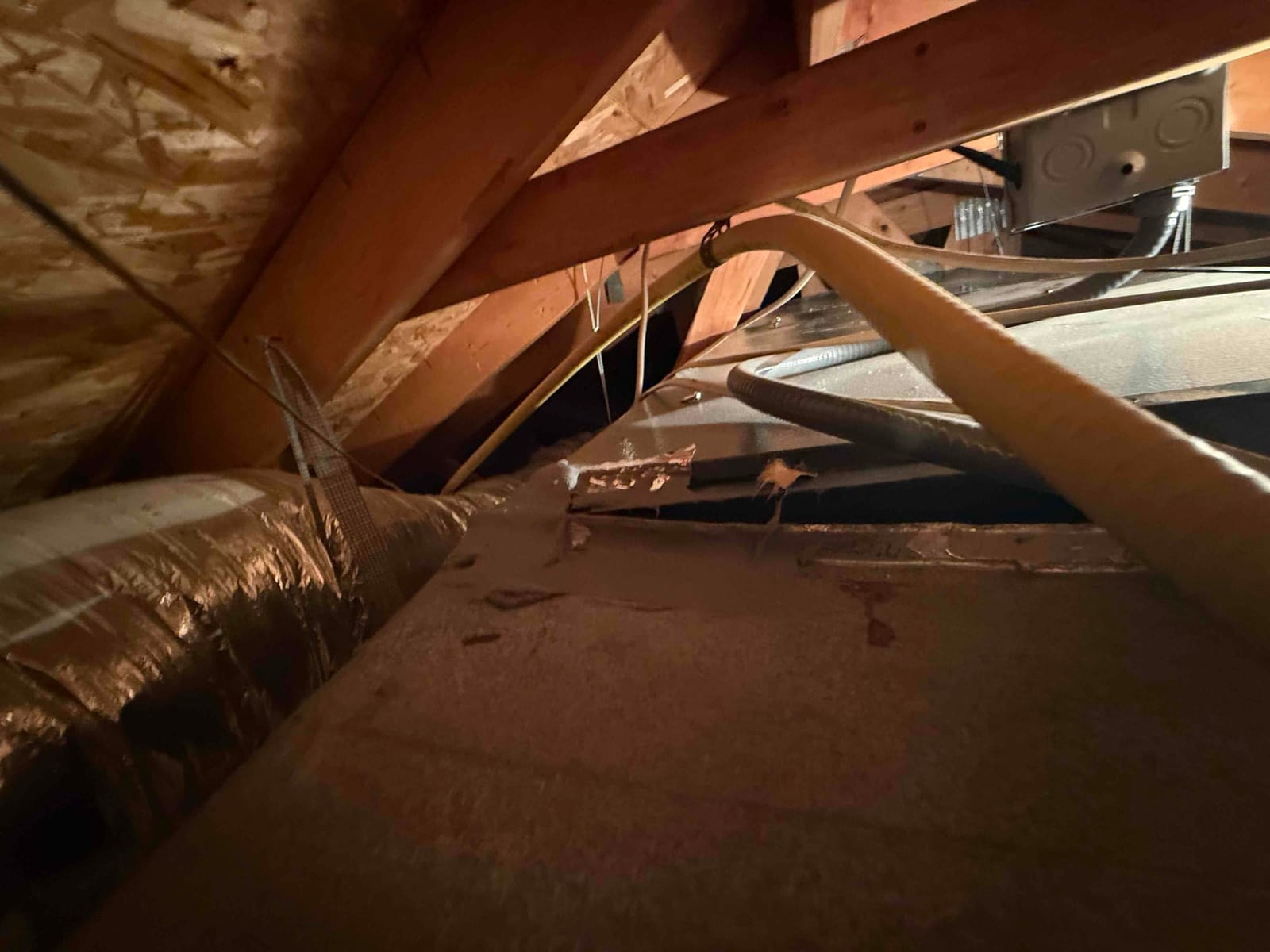 Attic air handler with insulated flex duct, electrical conduit, and condensate drain hose visible across dusty cabinet surface.