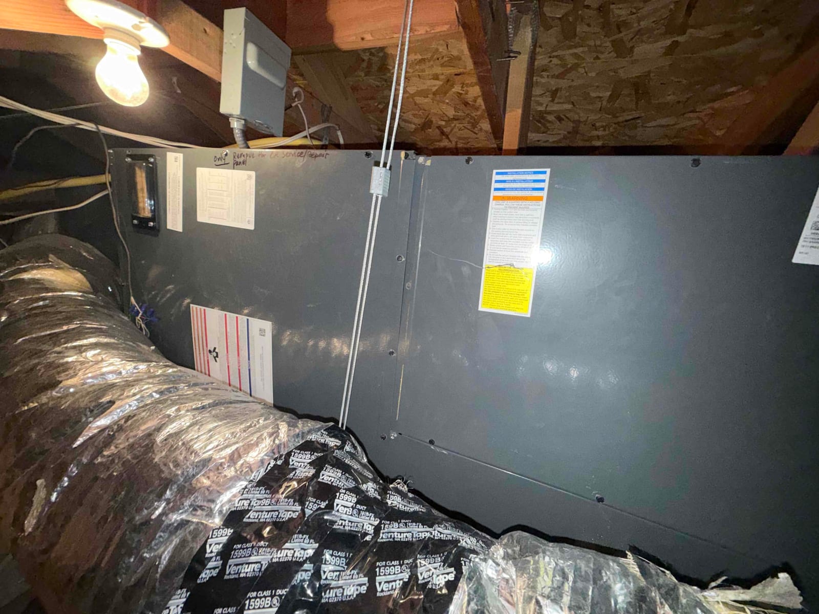 Attic-mounted air handler with insulated flexible duct attached, closed service panels, and multiple warning labels visible under a utility light.
