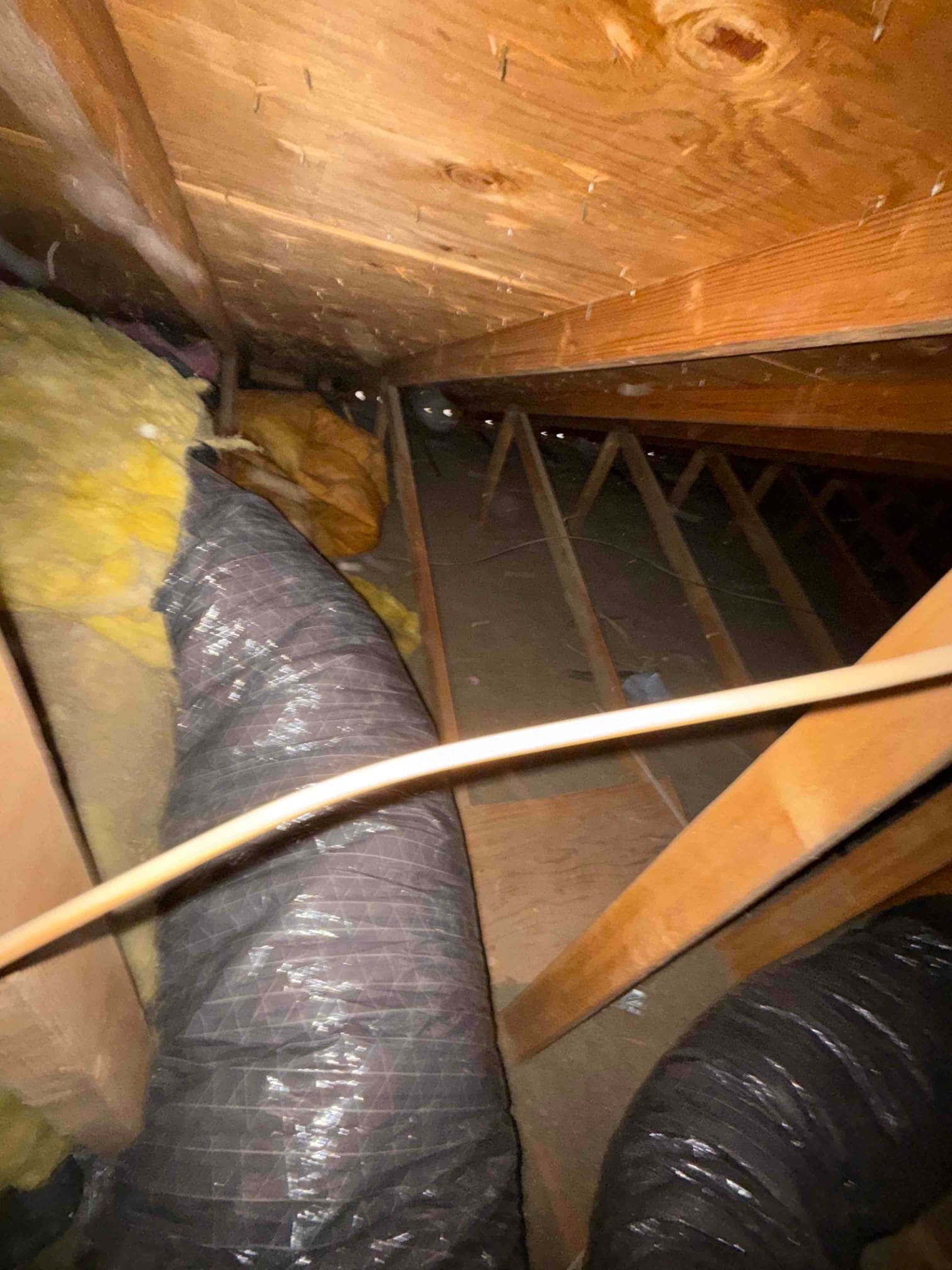 Attic space showing black flexible HVAC ducts routed between wooden framing with surrounding fiberglass insulation.