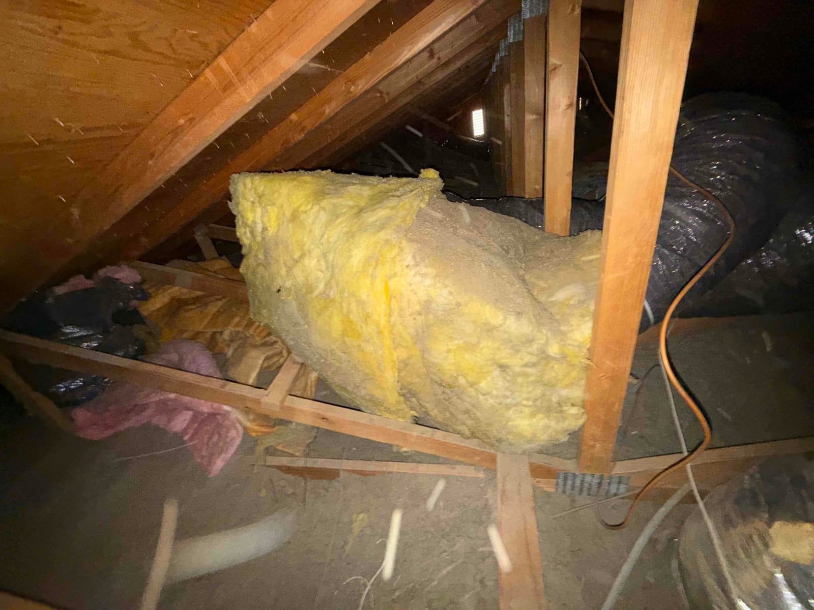 Attic space showing flexible HVAC ducting and a large piece of yellow fiberglass insulation resting over joists.