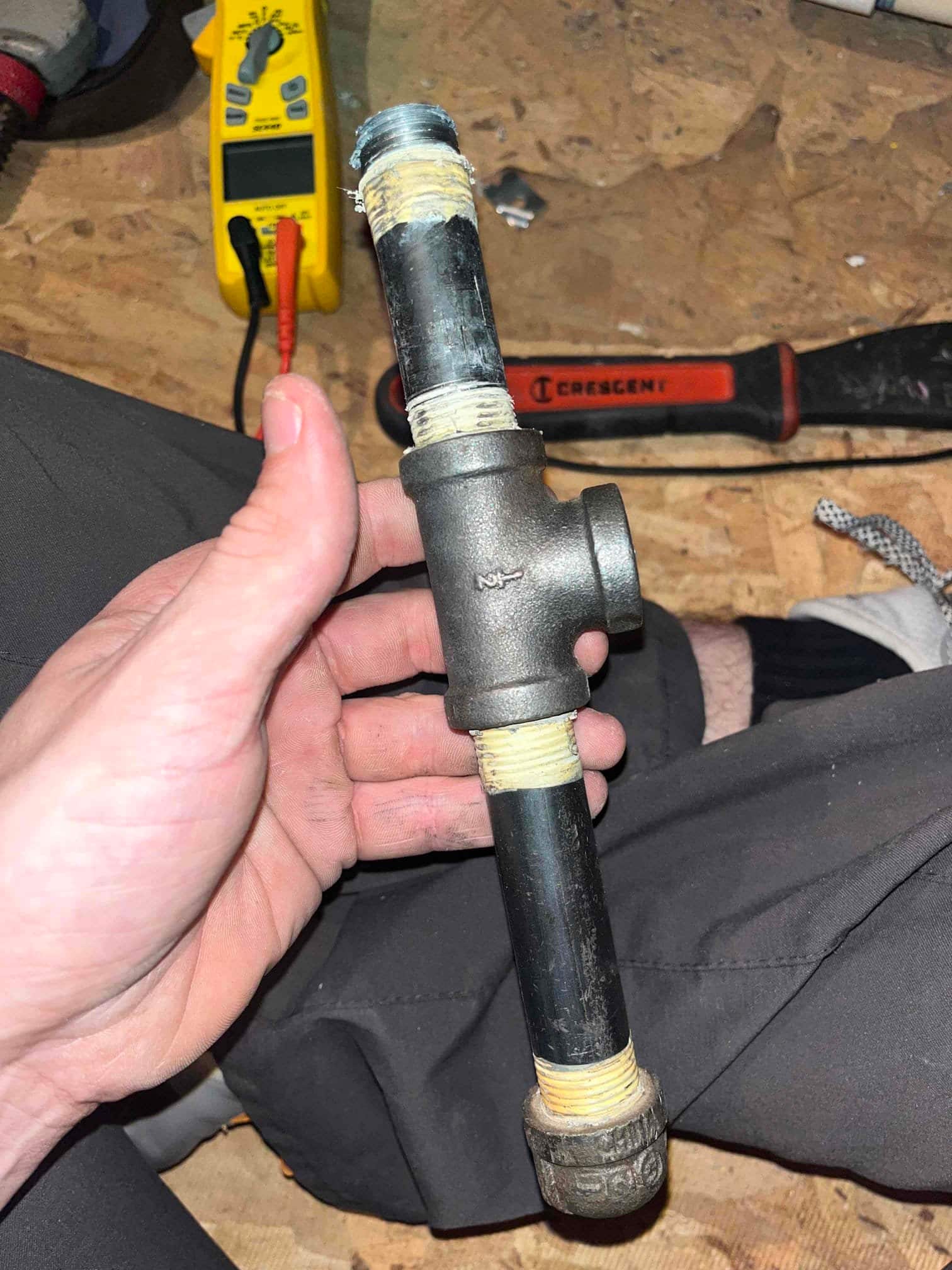 Black iron pipe assembly with T-fitting and threaded nipples, threads wrapped with sealant, held by technician on a work surface.