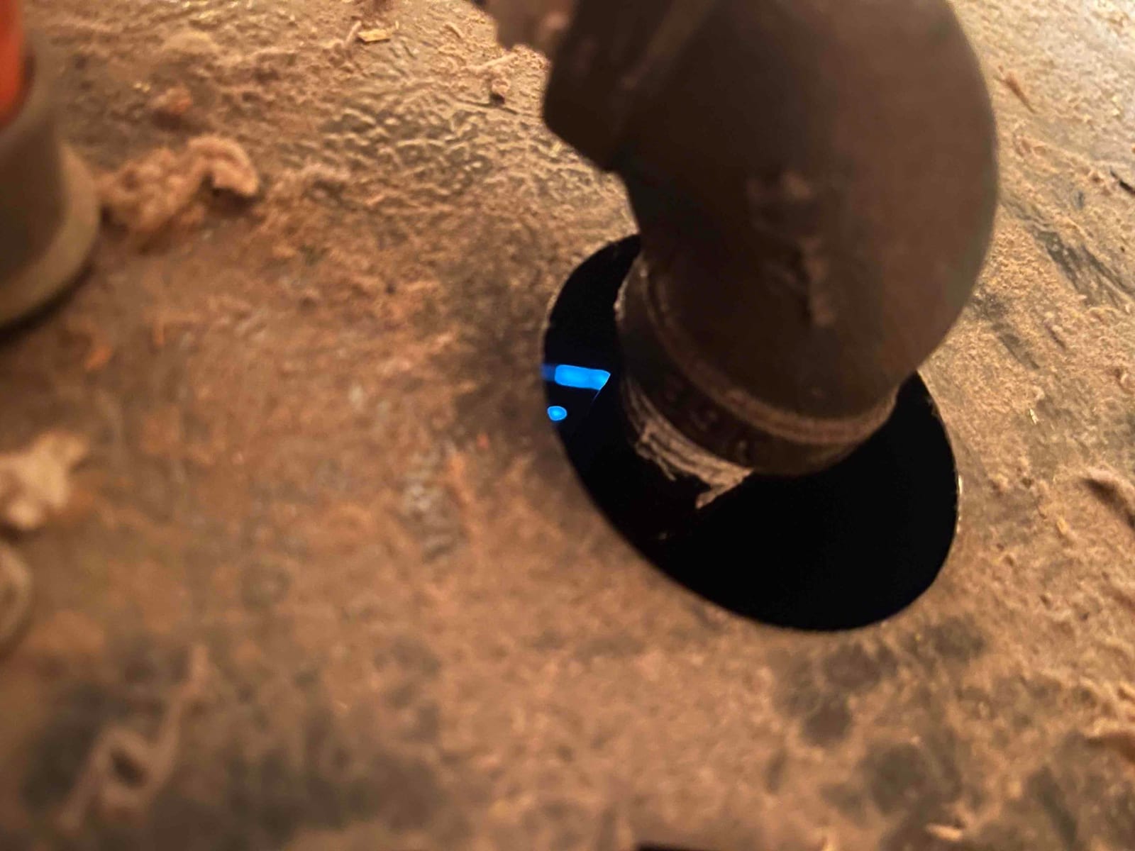 Blue burner flame is visible through a circular opening beside a threaded 90-degree elbow on a dusty metal appliance housing.
