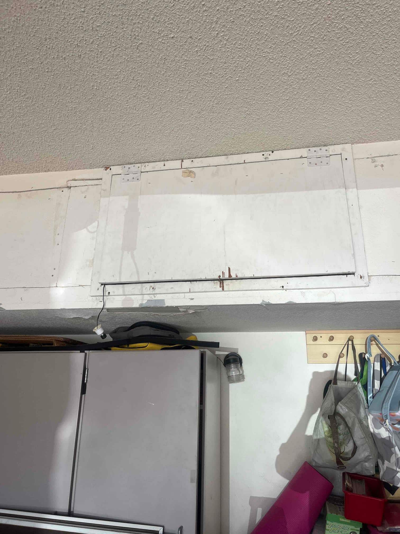 Ceiling access panel with metal hinges and rust stains above garage cabinets, with a hanging light socket and surrounding storage.
