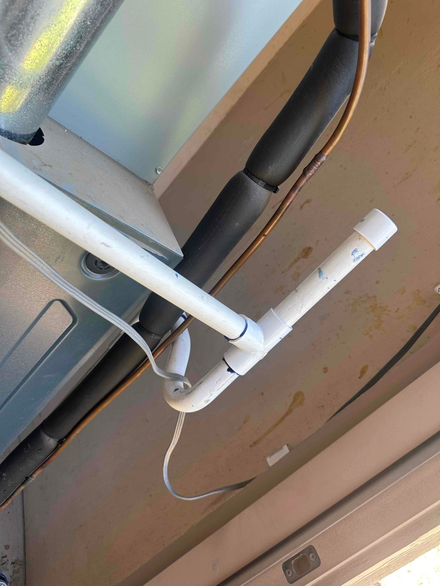 Condensate drain piping with cleanout tee and elbow beneath the air handler, alongside insulated refrigerant lines and copper tubing.