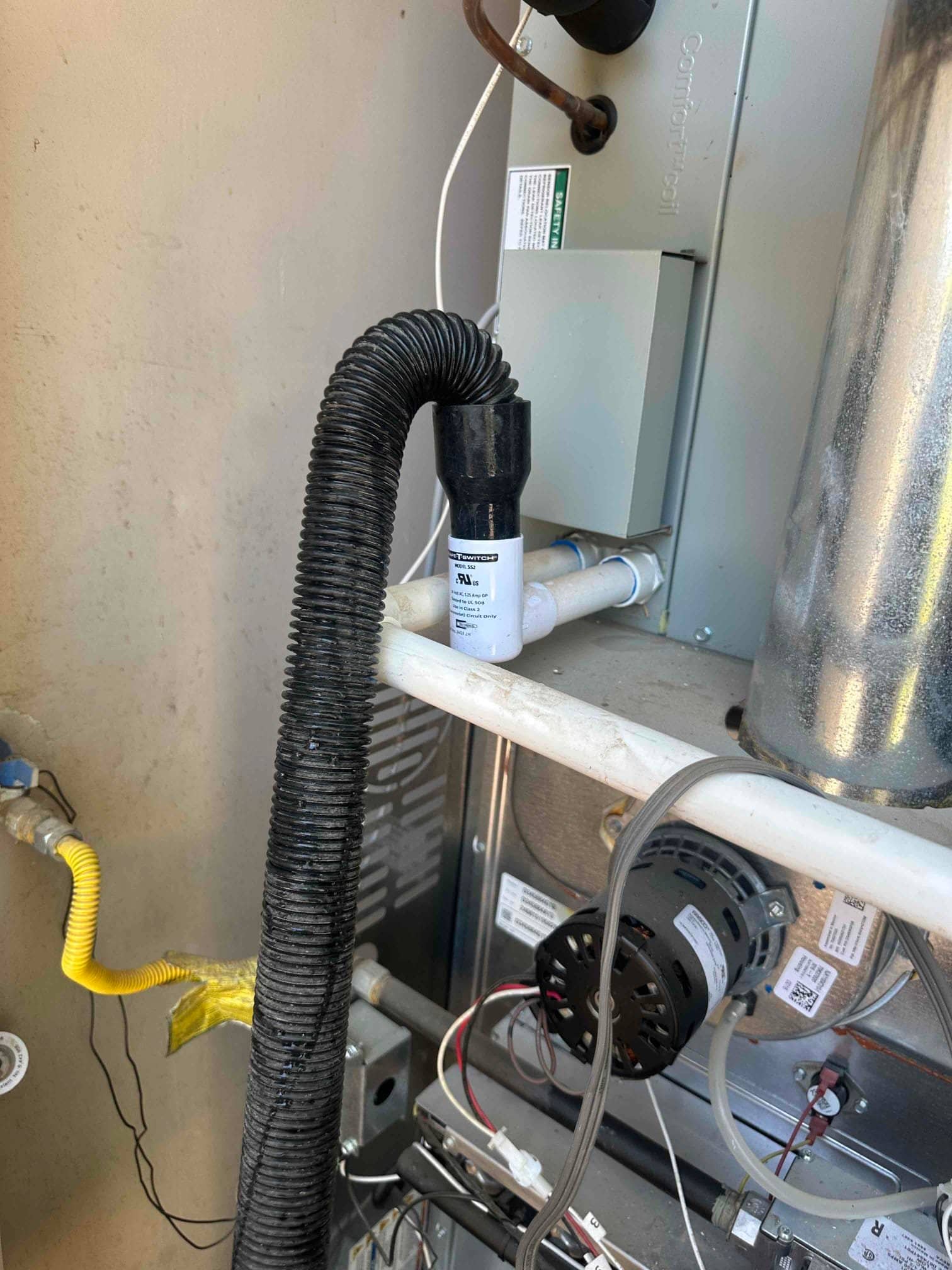 Condensate float switch installed on PVC drain line near furnace blower compartment with wiring and gas line visible.