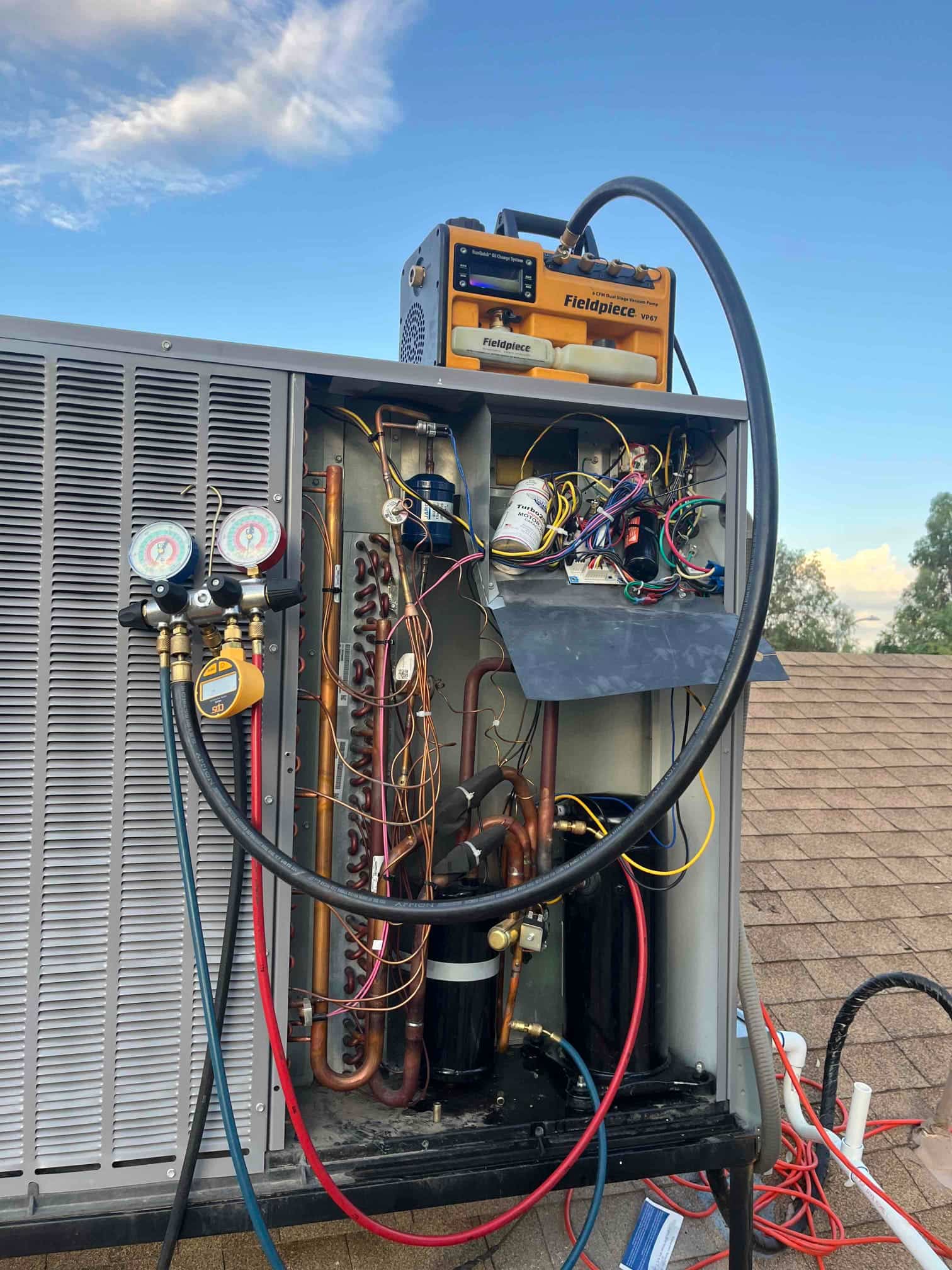 Condenser cabinet open, showing compressor, coil, copper tubing, and controls, with manifold gauges and a vacuum pump connected to the system.