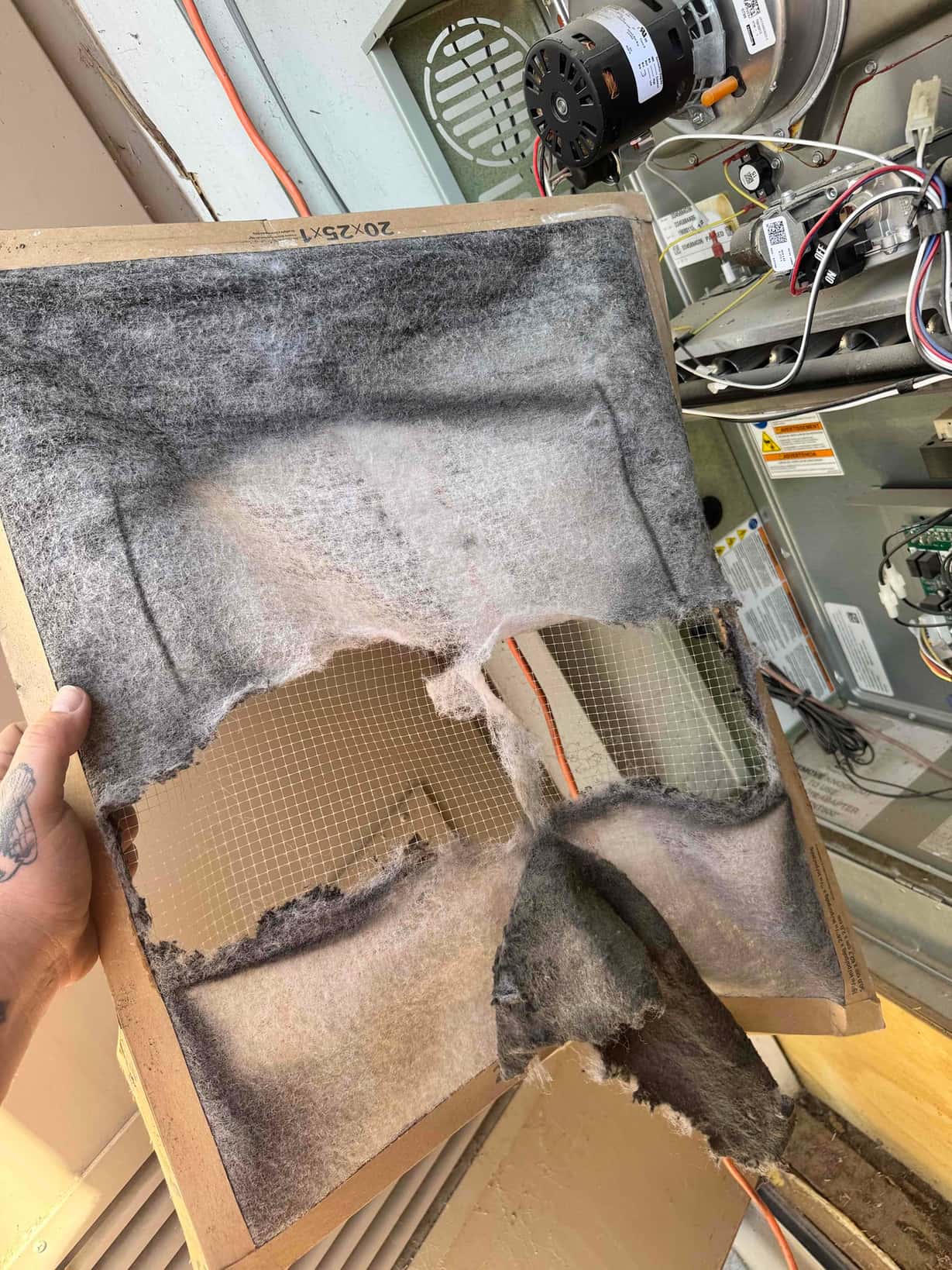 Damaged 20x25x1 fiberglass air filter with large tears, held in front of open furnace cabinet.