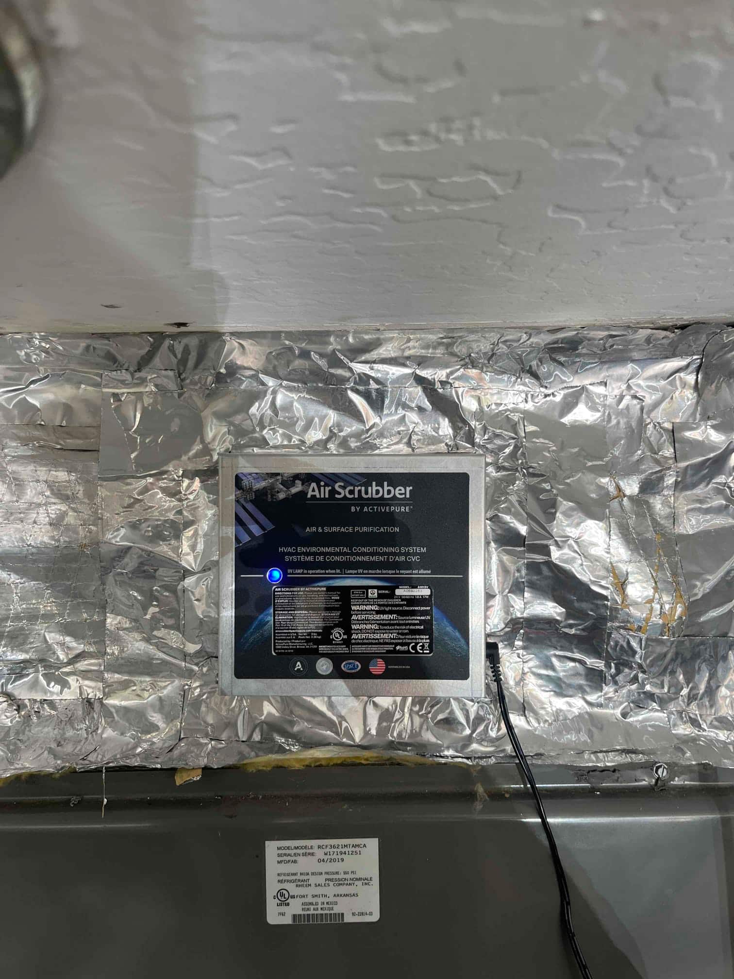 Duct-mounted air purification device installed on the supply plenum, with foil tape sealing and a low-voltage power cable connected.