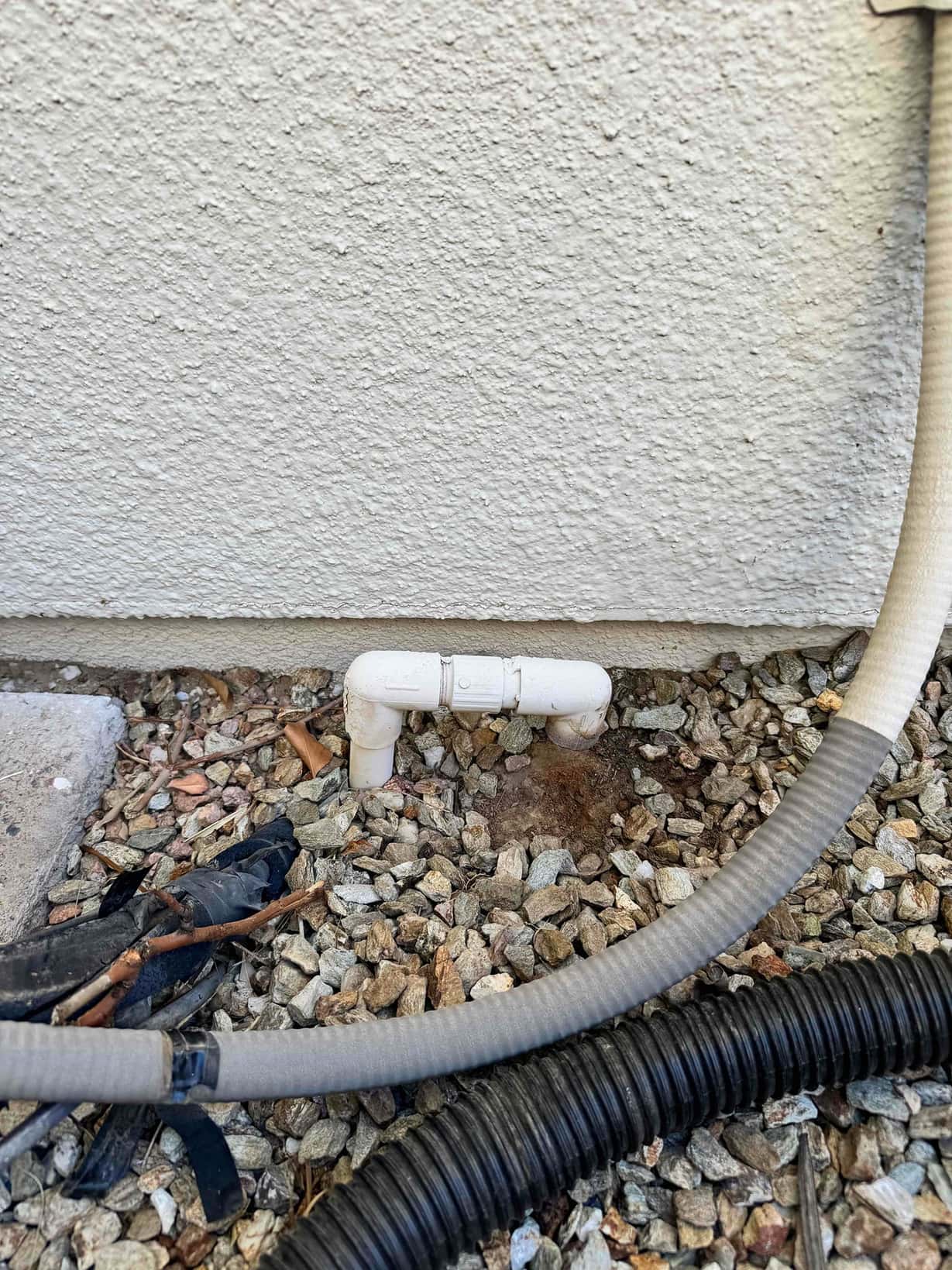 Exterior PVC condensate drain outlet at the wall, with cleanout tee and elbow above landscape gravel.