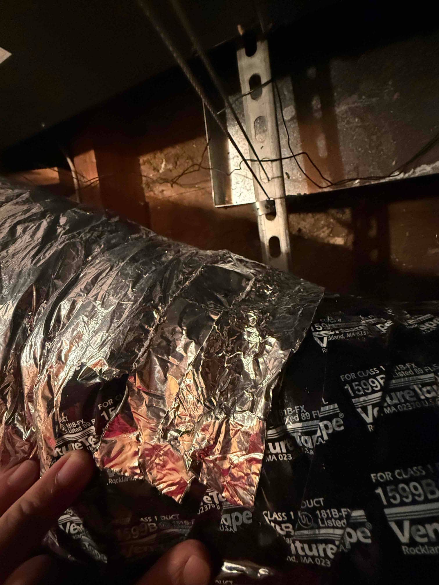Foil tape patched on flexible insulated duct near a metal support bracket, technician's fingers holding edge.