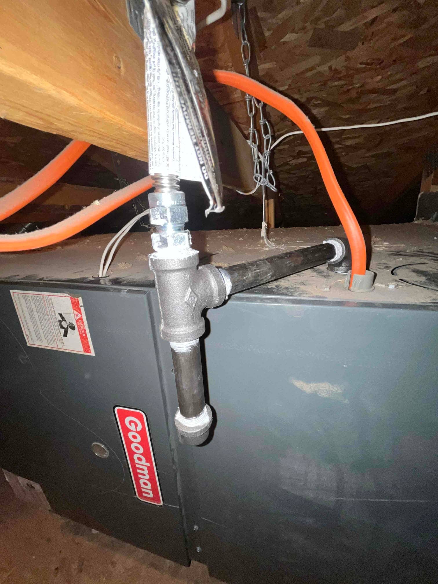 Gas furnace in attic with black iron gas piping and drip leg, flexible connector, and orange tubing above the cabinet.