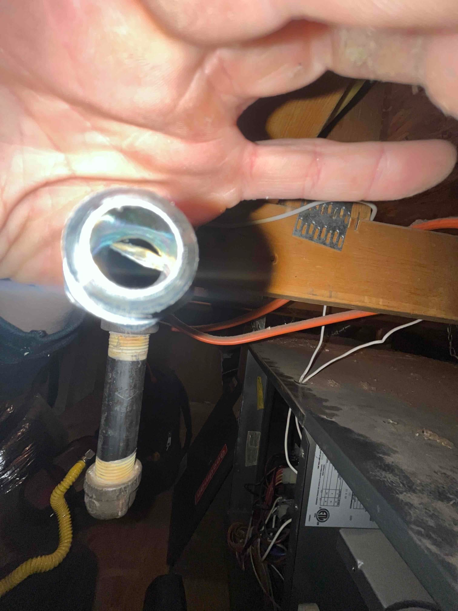 Hand holds an open metal fitting near the furnace cabinet, with gas piping, wiring, and service panel visible.