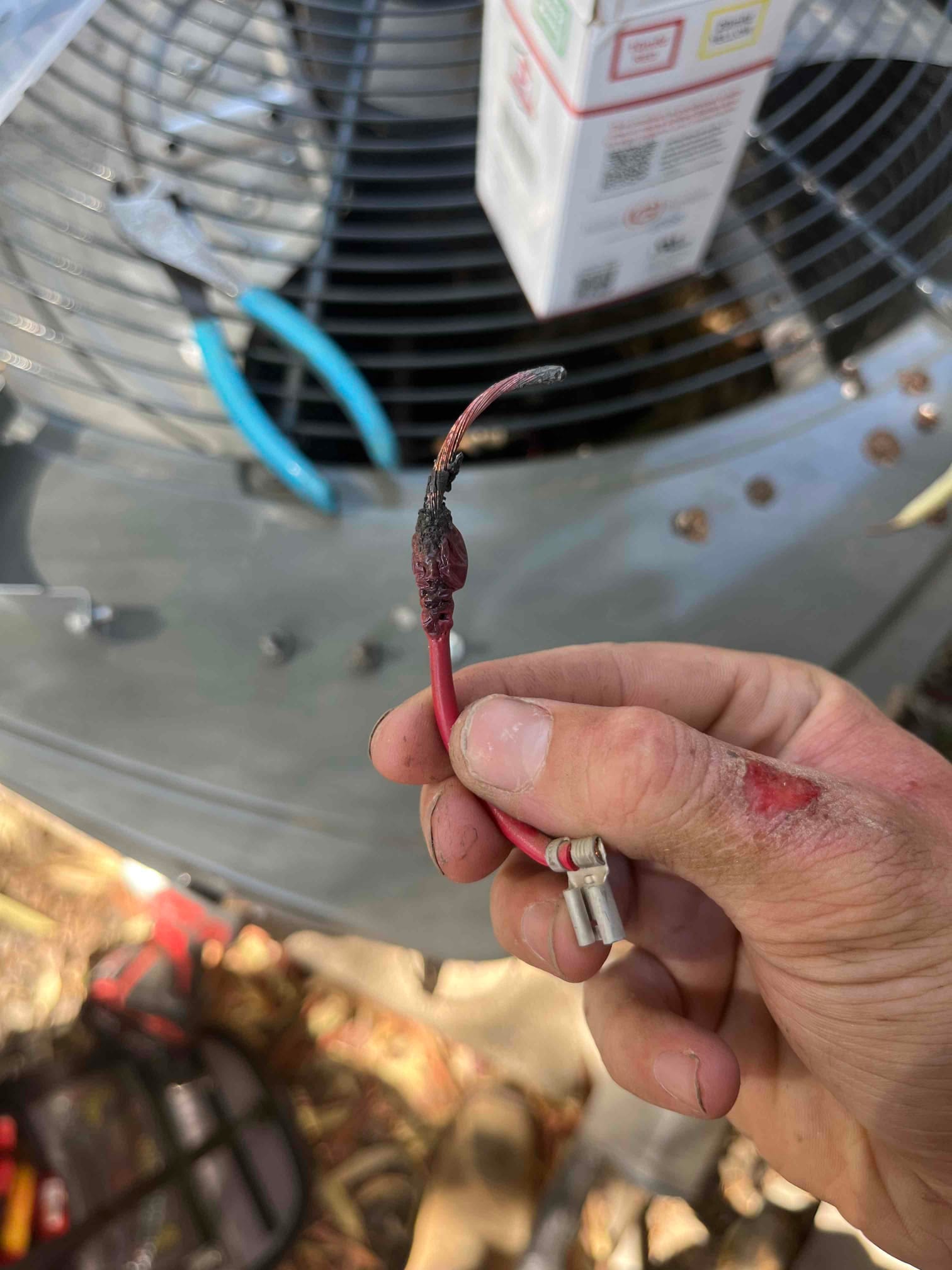 Hand holds a red electrical wire lead with charred insulation and exposed copper, in front of an outdoor condenser fan.
